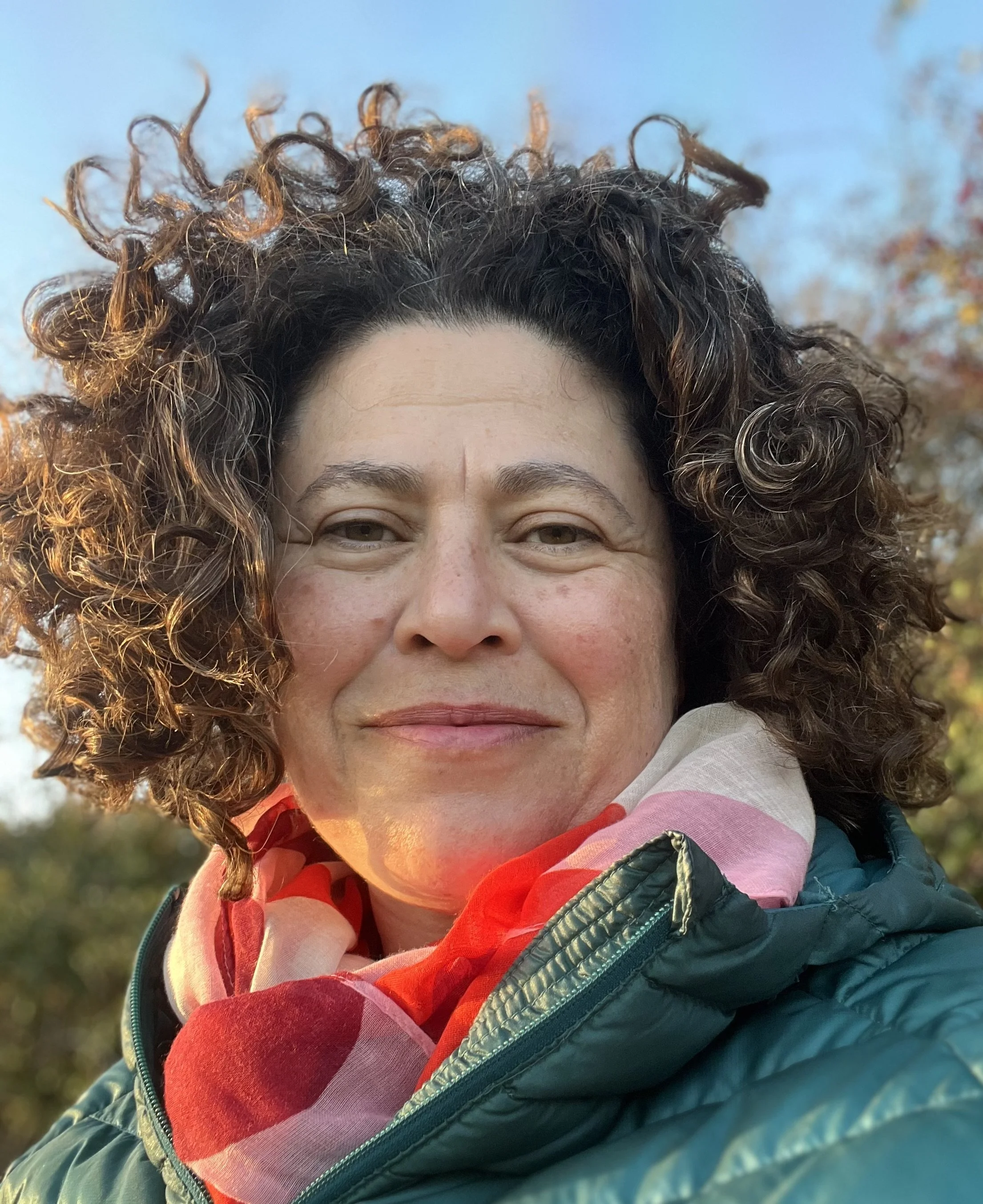 Close-up of a woman with curly brown hair, smiling outdoors, wearing a colorful scarf and a quilted jacket.