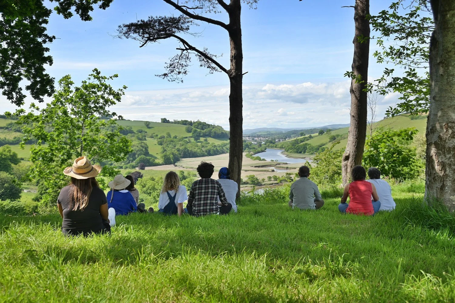 Group of people sitting on green grass under trees, overlooking a scenic landscape of rolling hills, trees, and a winding river under a partly cloudy sky.
