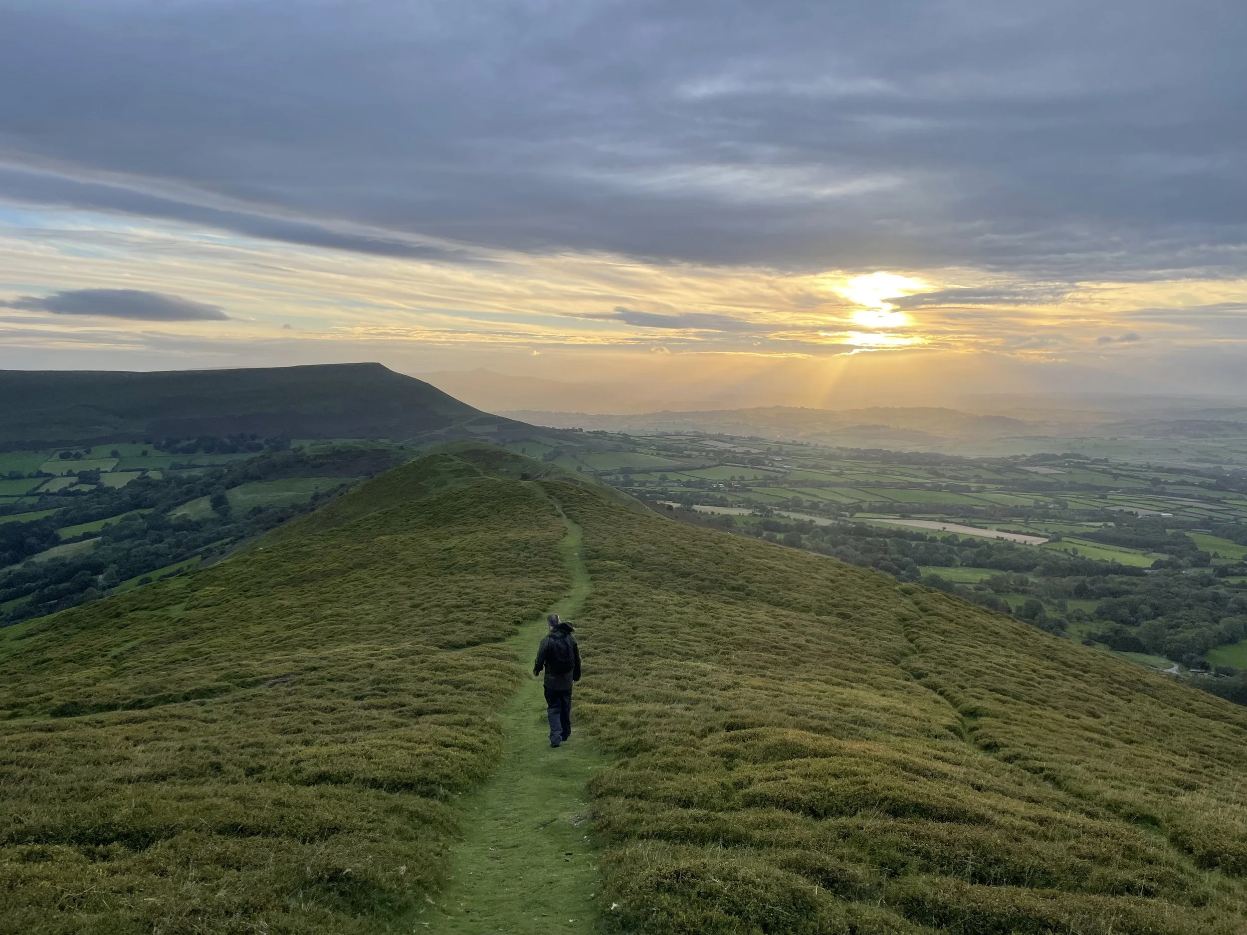 A person walking along a trail on a green hillside during sunset with a cloudy sky and distant landscape.