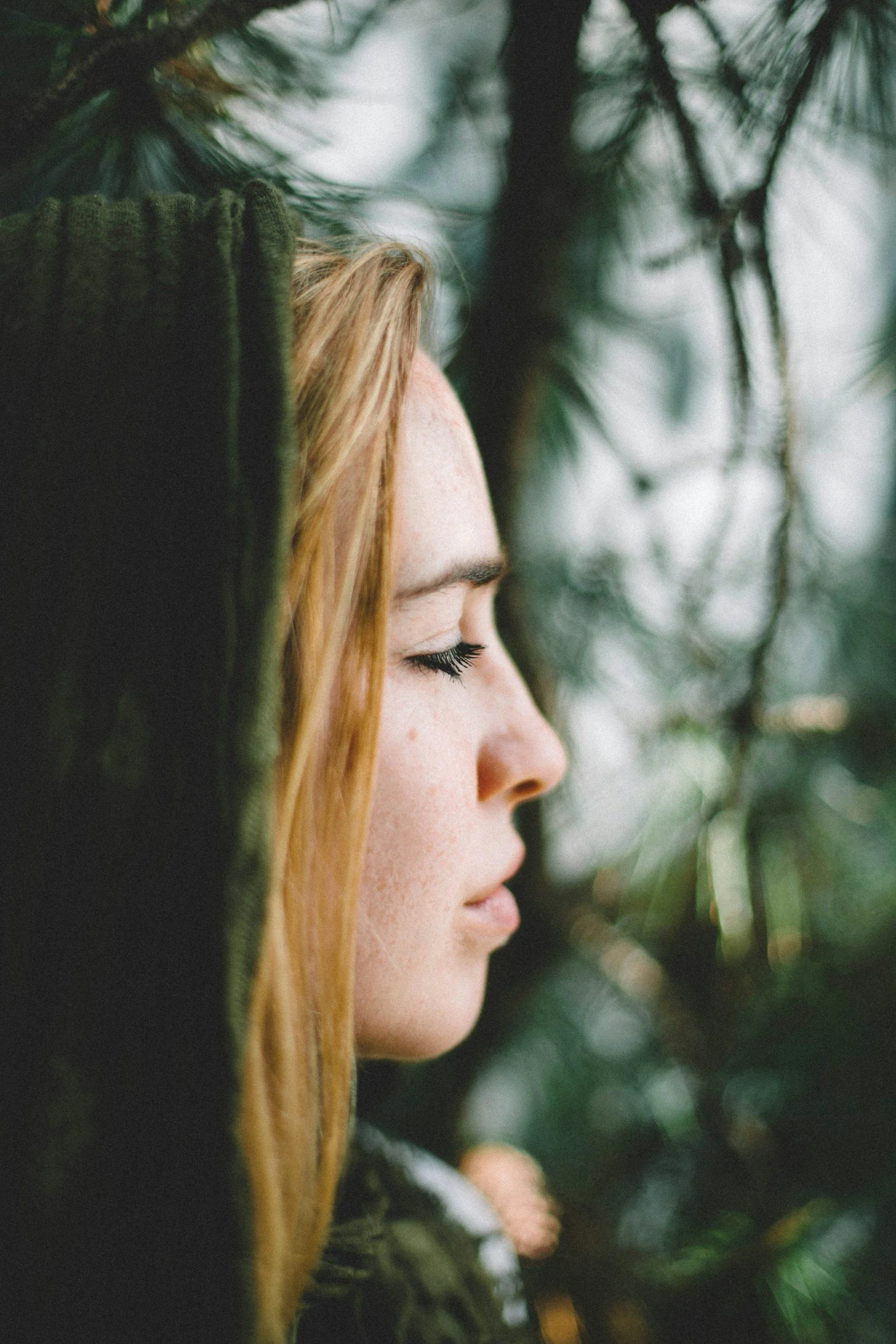Side profile of a young woman with closed eyes, red hair, and freckles, surrounded by green pine branches.