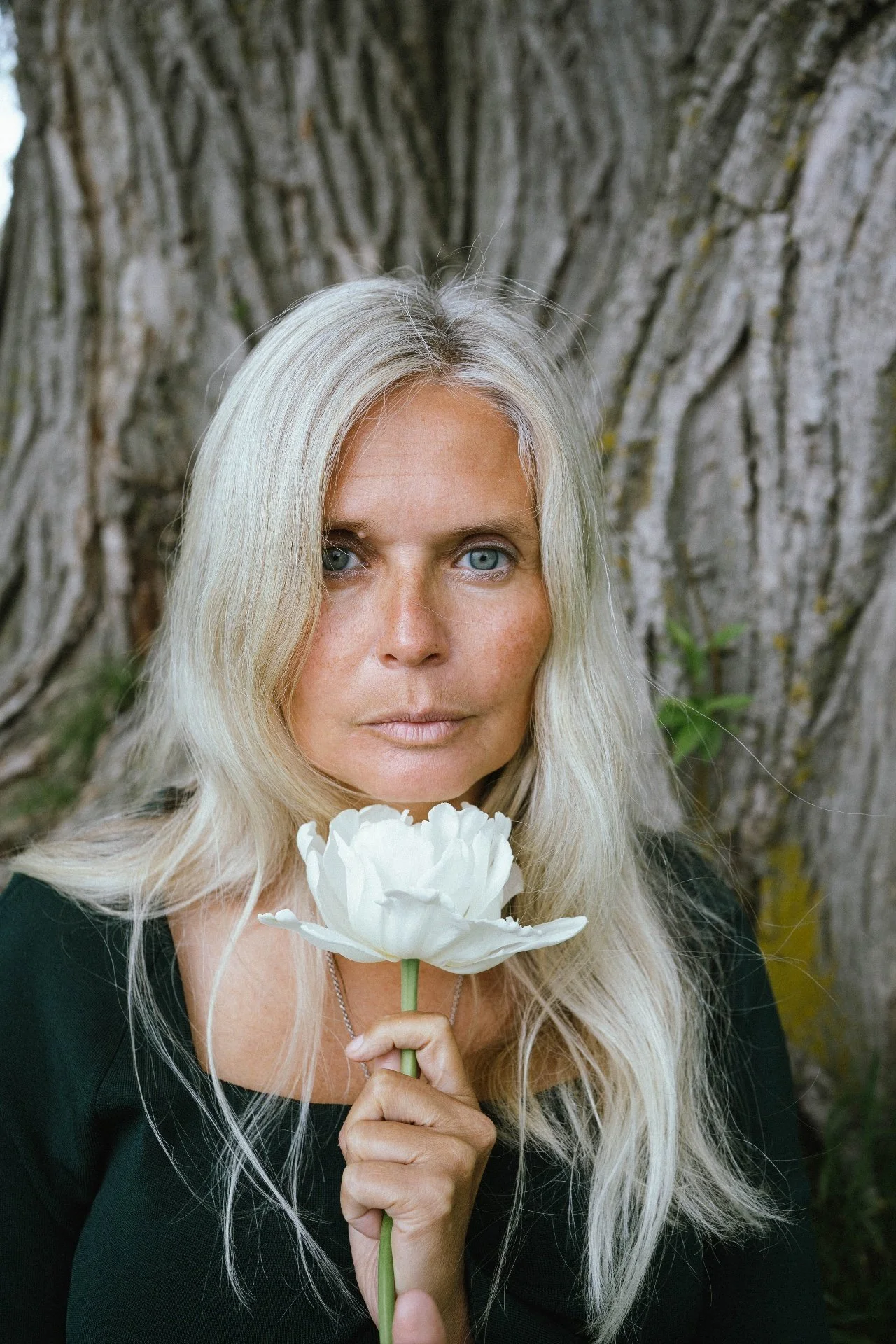 A woman with long blonde hair and blue eyes holding a white peony flower close to her face, standing outdoors in front of a large tree trunk.