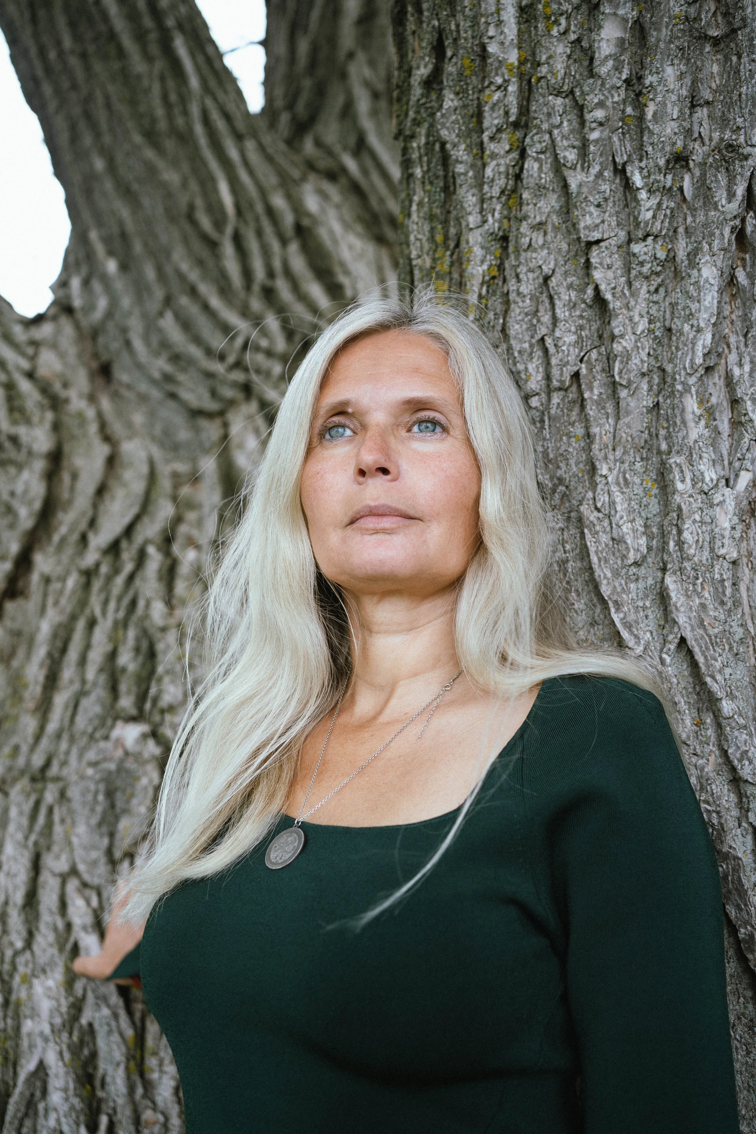 A woman with long blonde hair and blue eyes leaning against a large tree trunk outdoors.