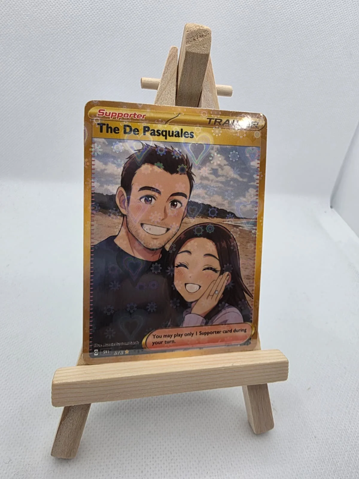 A trading card featuring a cartoon illustration of a smiling man and woman, with the man winking. The card is displayed on a small wooden easel. The background of the card has a landscape with clouds and trees, and there are floral and heart-shaped p
