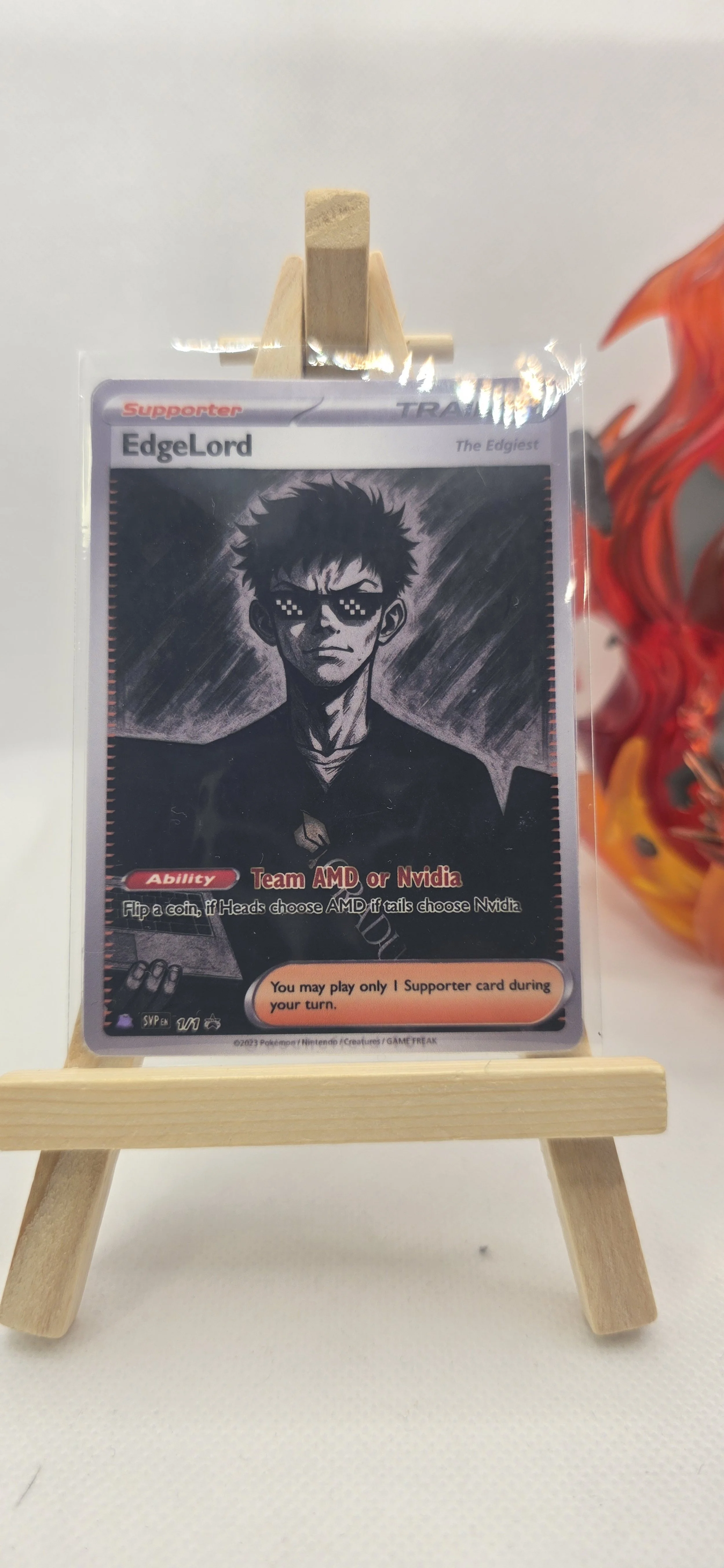 A Pokémon trading card titled 'EdgeLord' with a black and white illustration of a male character wearing pixelated sunglasses and a dark outfit, displayed on a small wooden easel, with a red and orange fire sculpture on the right side of the image.