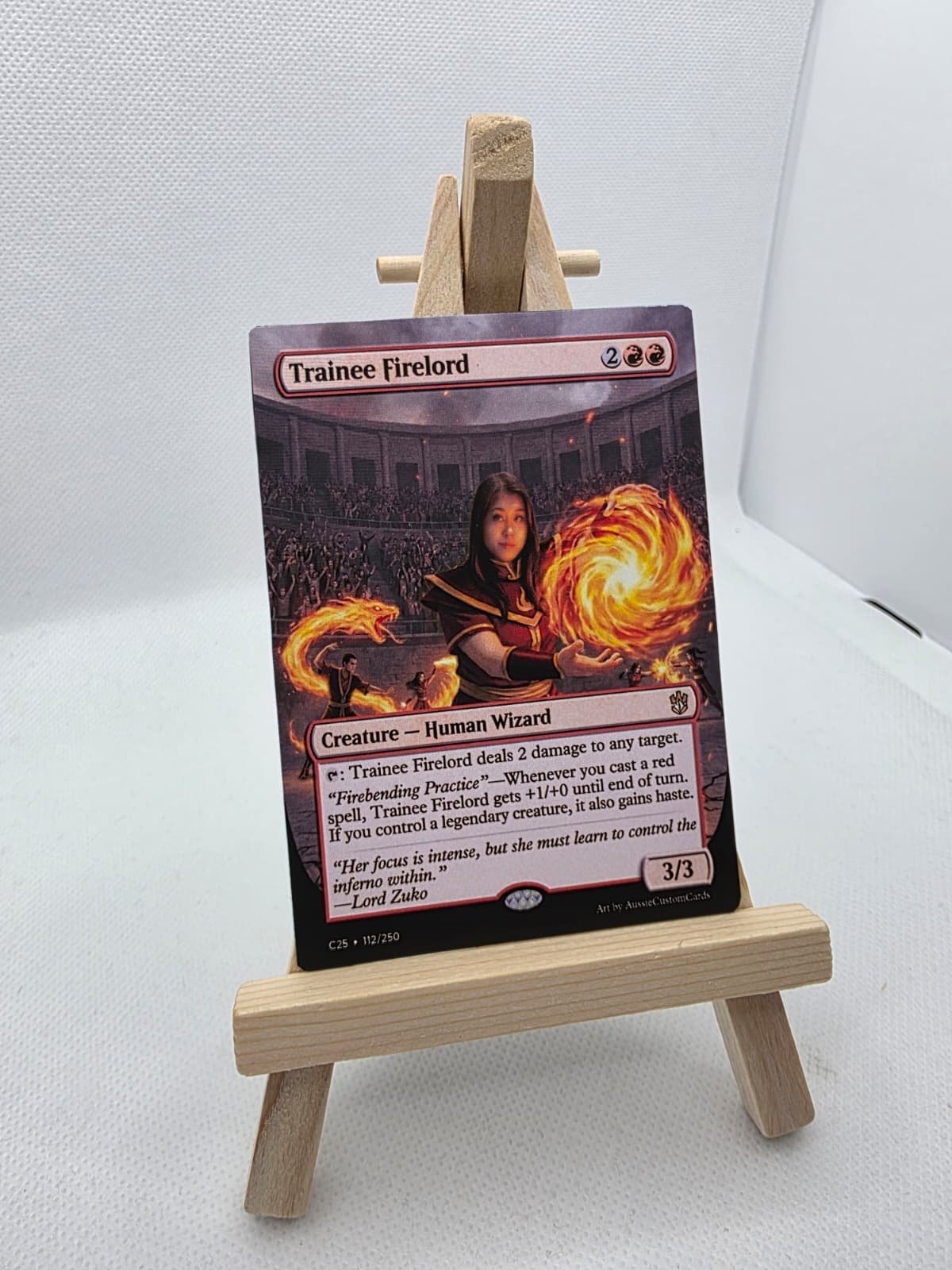 A custom Magic: The Gathering card named Trainee Firelord displayed on a small wooden easel. The card features a female human wizard casting fire spells in an arena setting.