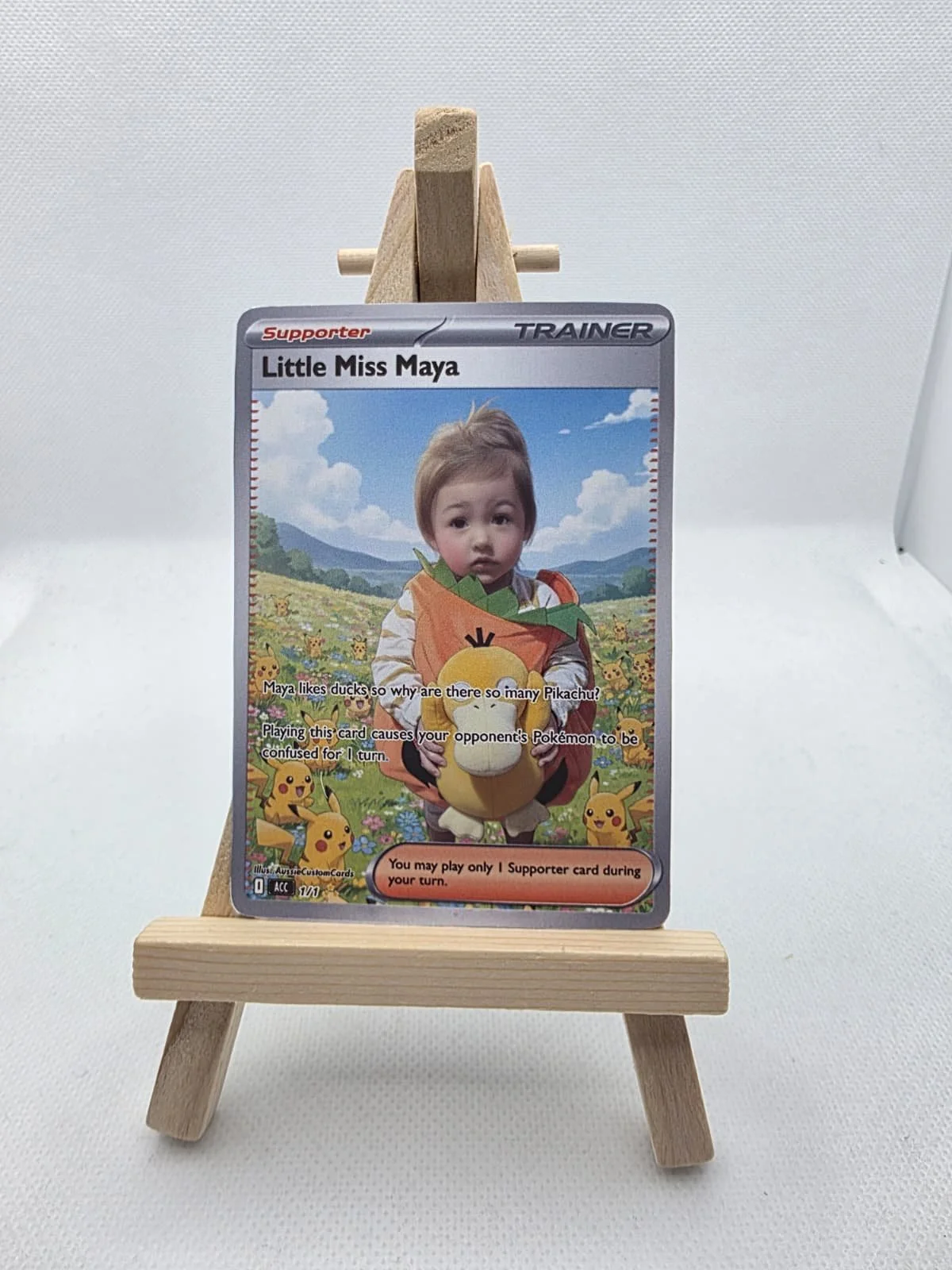 Custom Pokemon-themed supporter trainer card featuring a young child with short blonde hair, surrounded by Pikachu graphics, displayed on a small wooden easel against a plain white background.