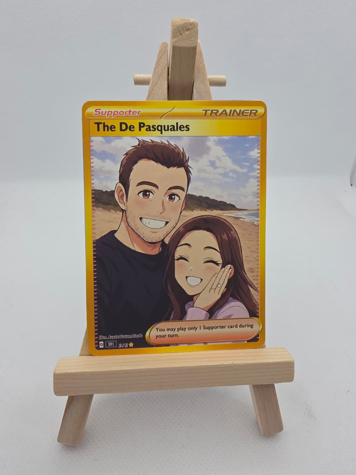 A Pokémon playing card featuring a cartoon couple smiling on a beach, with a small wooden easel displaying the card.
Anniversary present, love, wedding, present idea