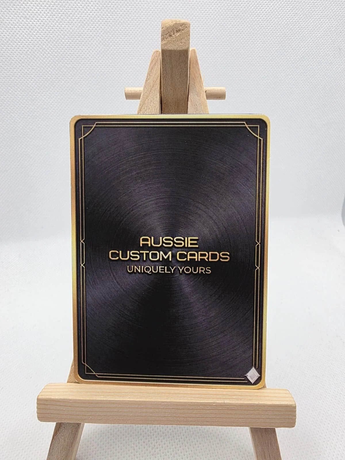 Black card with gold border and text that reads "Aussie Custom Cards, Uniquely Yours," displayed on a small wooden easel.