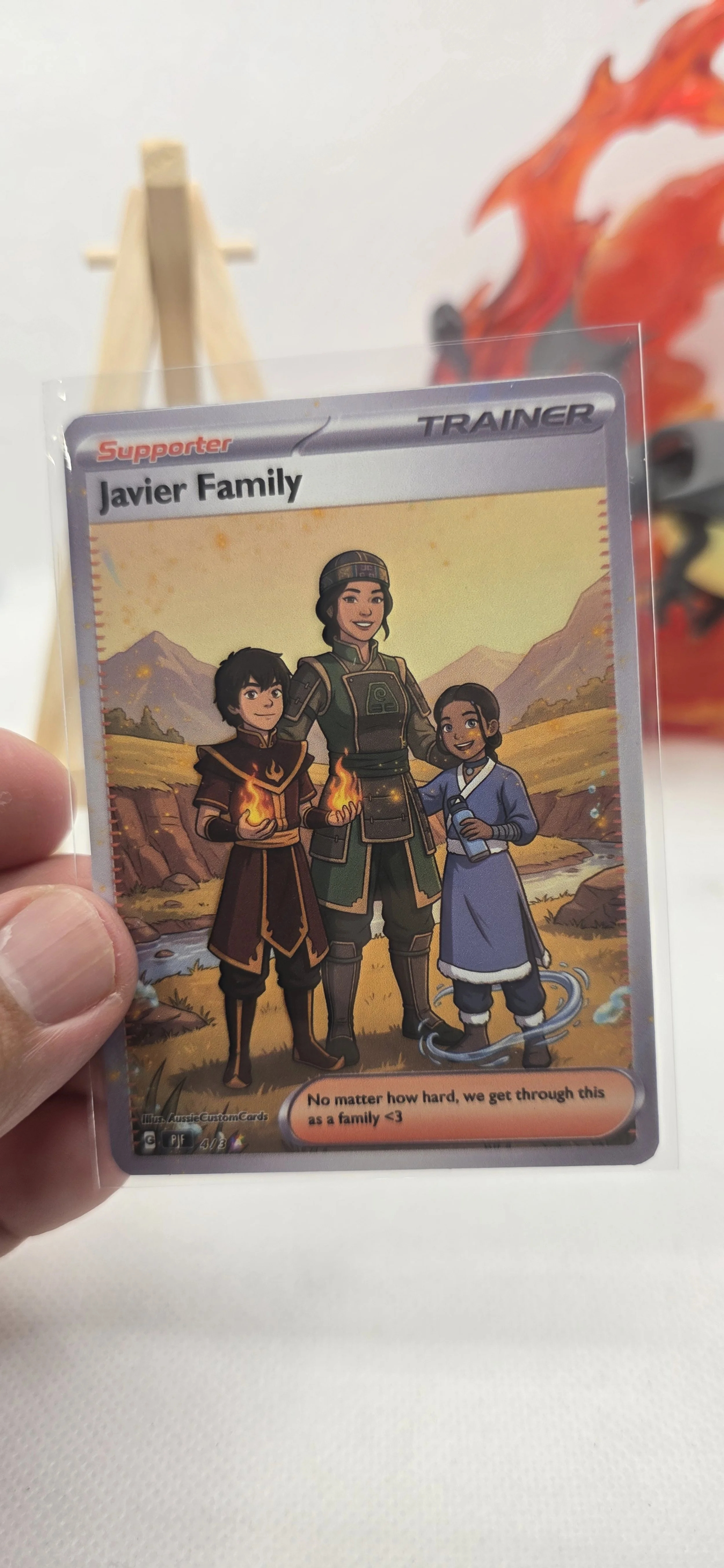 Illustrated supporter trading card featuring the Javier Family, with two children and an adult, in a natural outdoor setting with mountains in the background.