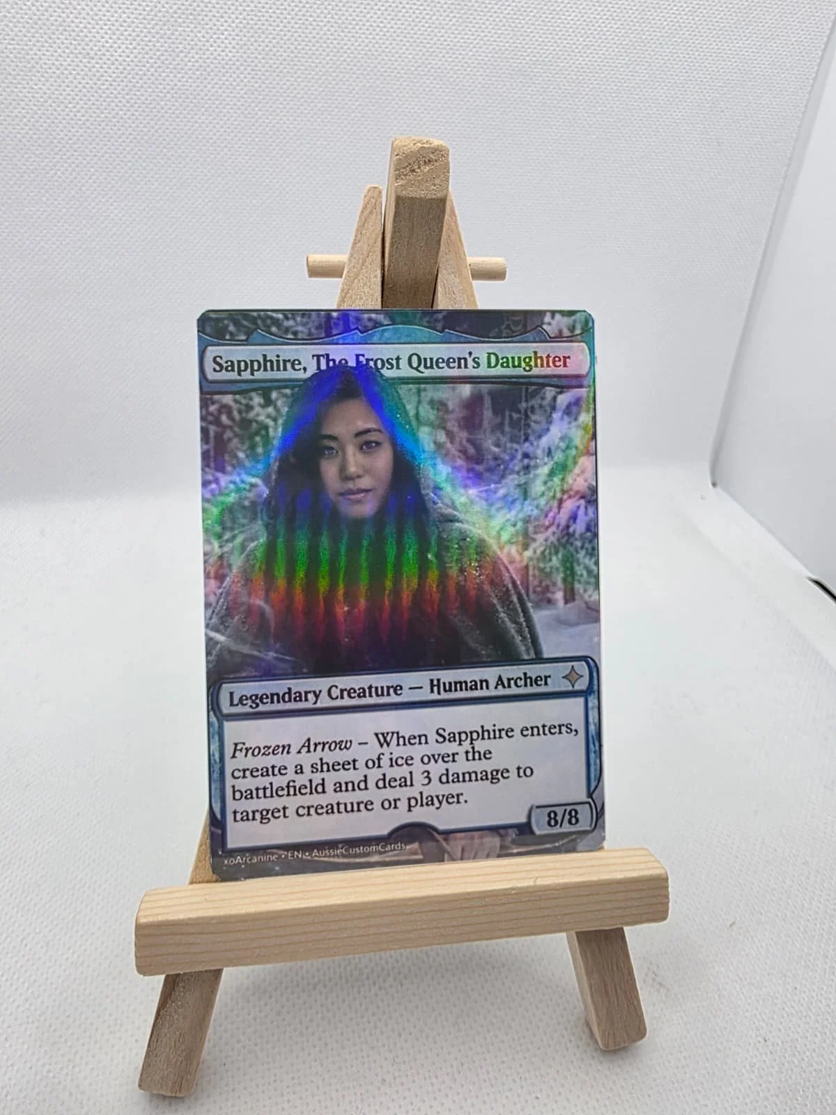 A holographic custom Magic: The Gathering card titled 'Sapphire, The Frost Queen's Daughter,' displayed on a small wooden easel, with a woman on the card's artwork and icy, colorful background.
