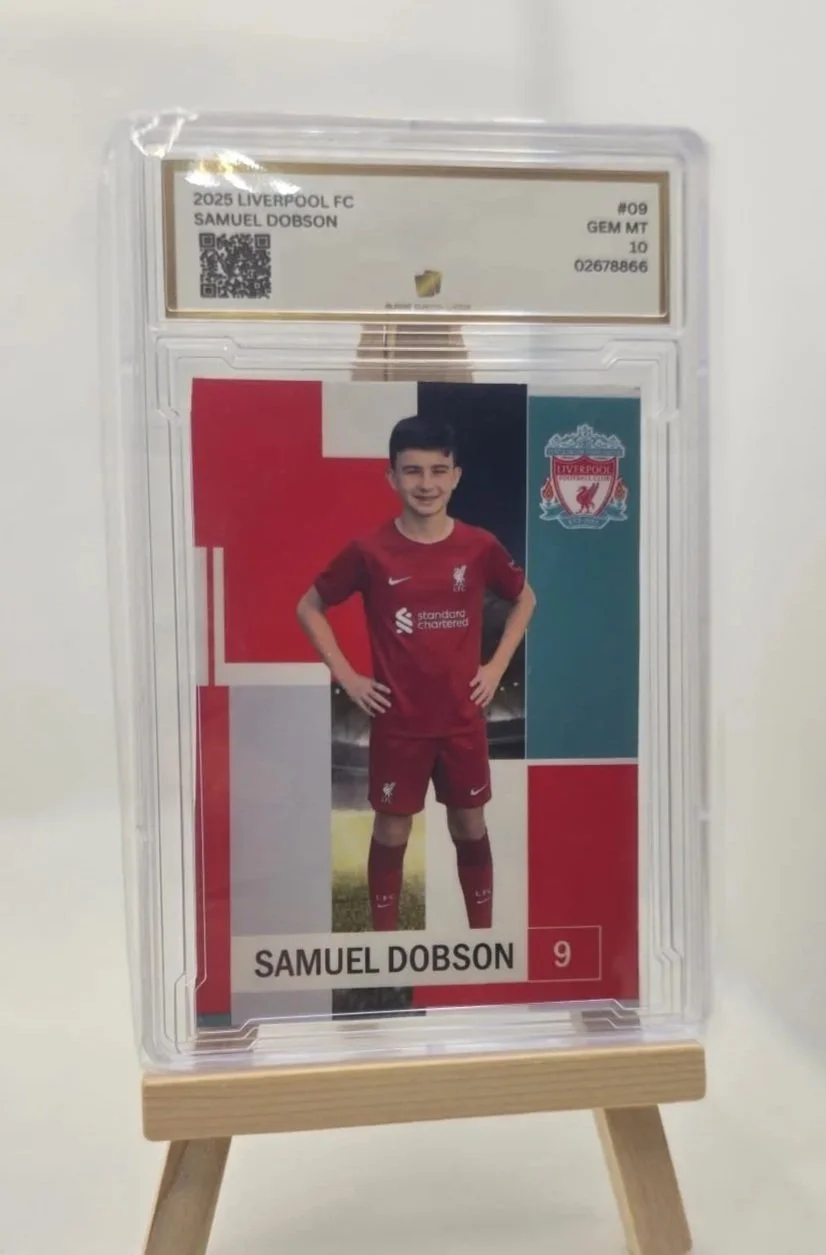 A graded soccer trading card of Samuel Dobson in a Liverpool FC kit, displayed on a small wooden easel.
