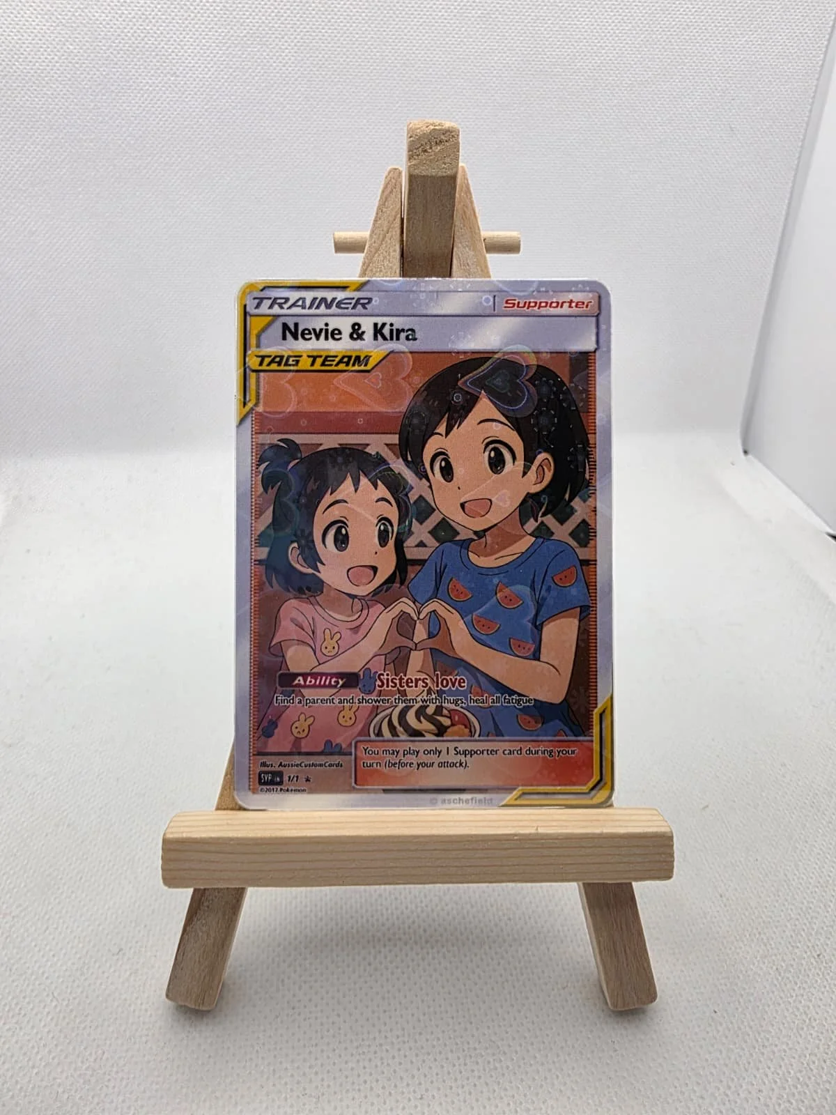 A Pokémon trading card titled 'Nevie & Kira' featuring two animated girls, one younger and the other older, making a heart shape with their hands. The card is in a clear plastic sleeve and stands on a small wooden easel against a white background.