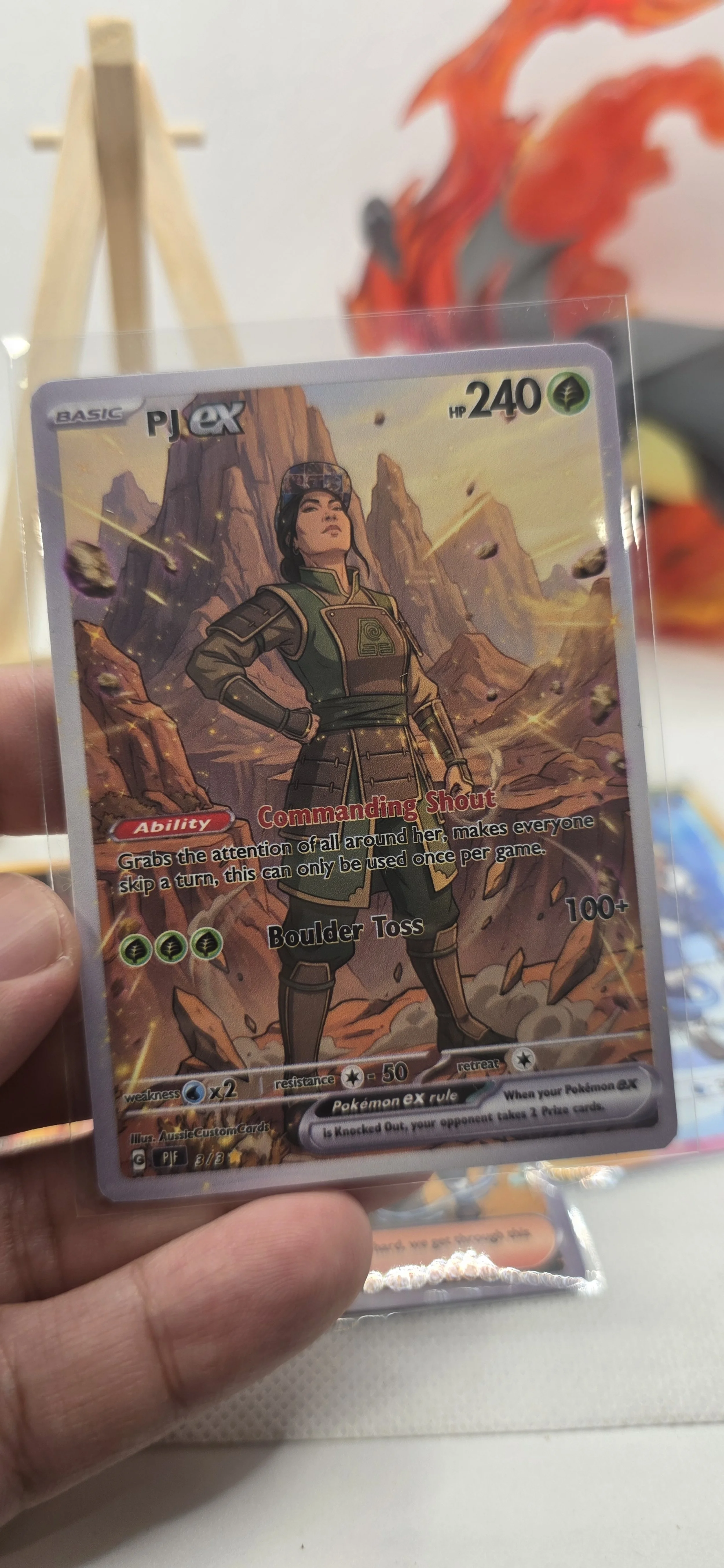 A Pokémon trading card featuring a character standing with hands on hips in front of a rocky desert landscape with mountains in the background. The card shows the character's name, 'PJ ex,' with 240 HP and various attack details.