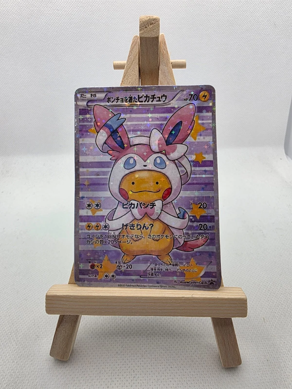 A Pokémon card featuring a character with large bunny ears, colorful accents, and a yellow face in a costume with pink and blue details. The card is displayed on a small wooden easel against a white background.
