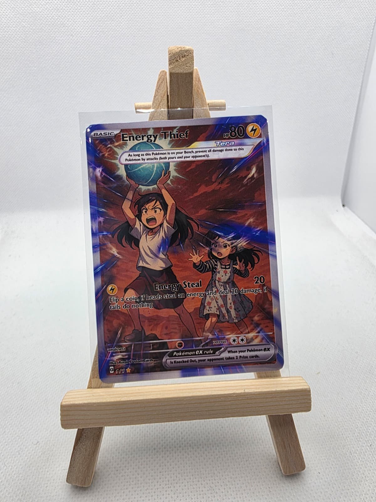 A Pokémon card titled 'Energy Thief' displayed on a small wooden stand. The card features animated characters, a girl holding a globe above her head, and another girl with long black hair, set against a fiery background.