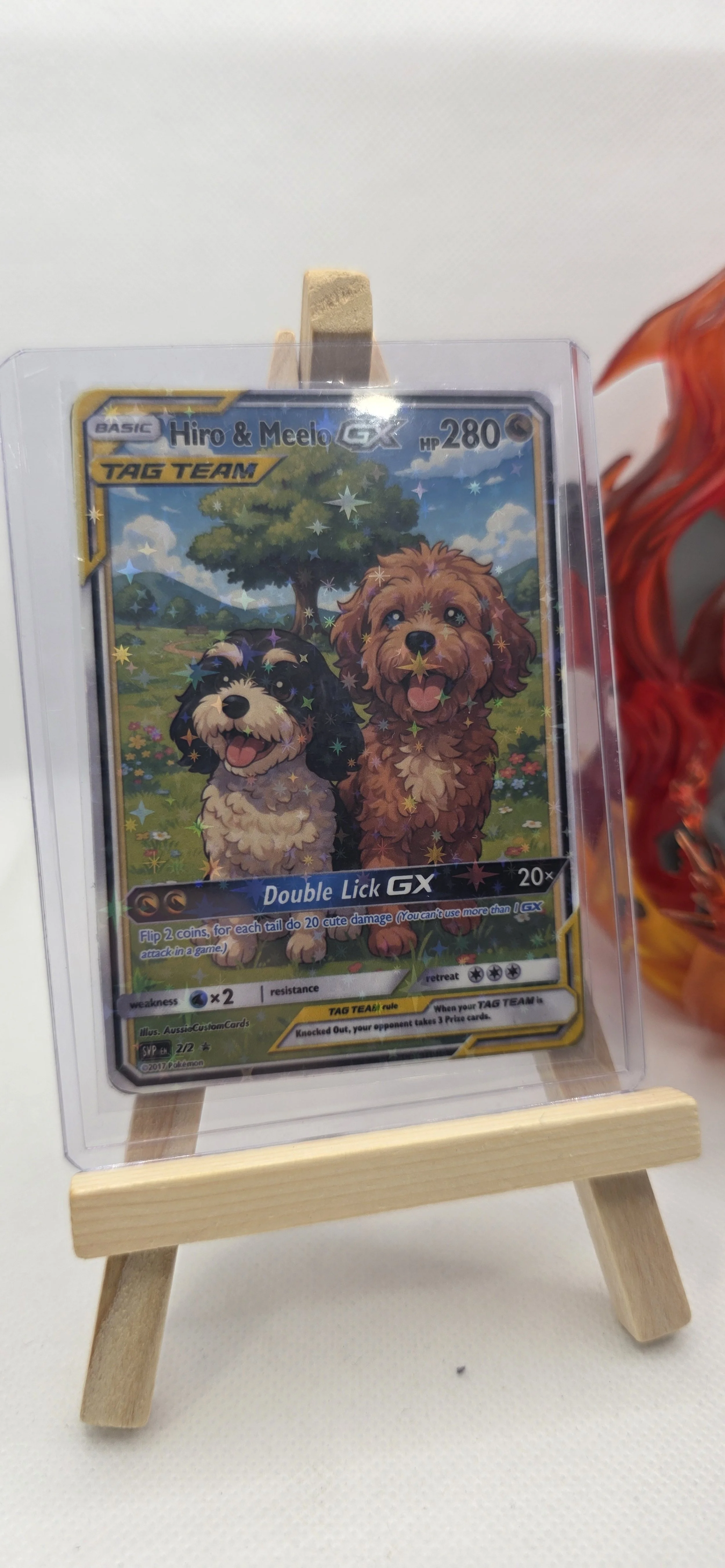 A Pokémon trading card titled 'Hiro & Meelo GX' featuring two cartoon dogs, set against a background of trees and a blue sky with clouds, displayed on a small wooden easel.