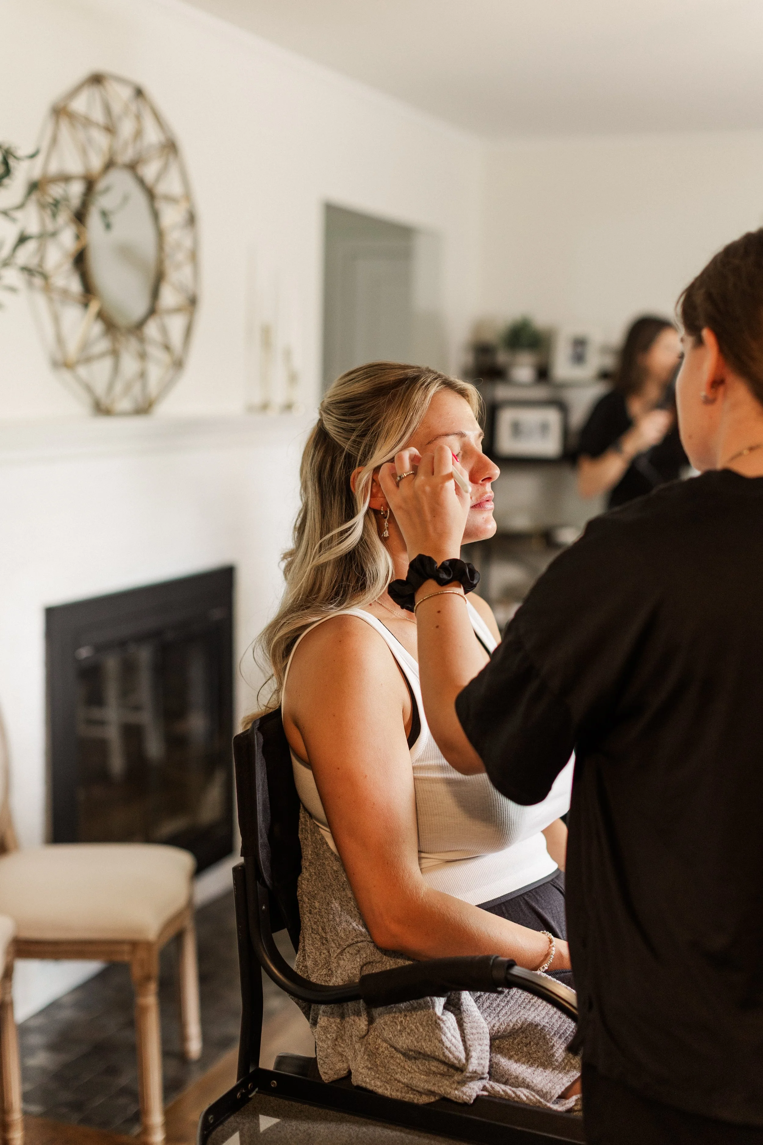Woman sitting in a chair having makeup applied by another person in a living room.