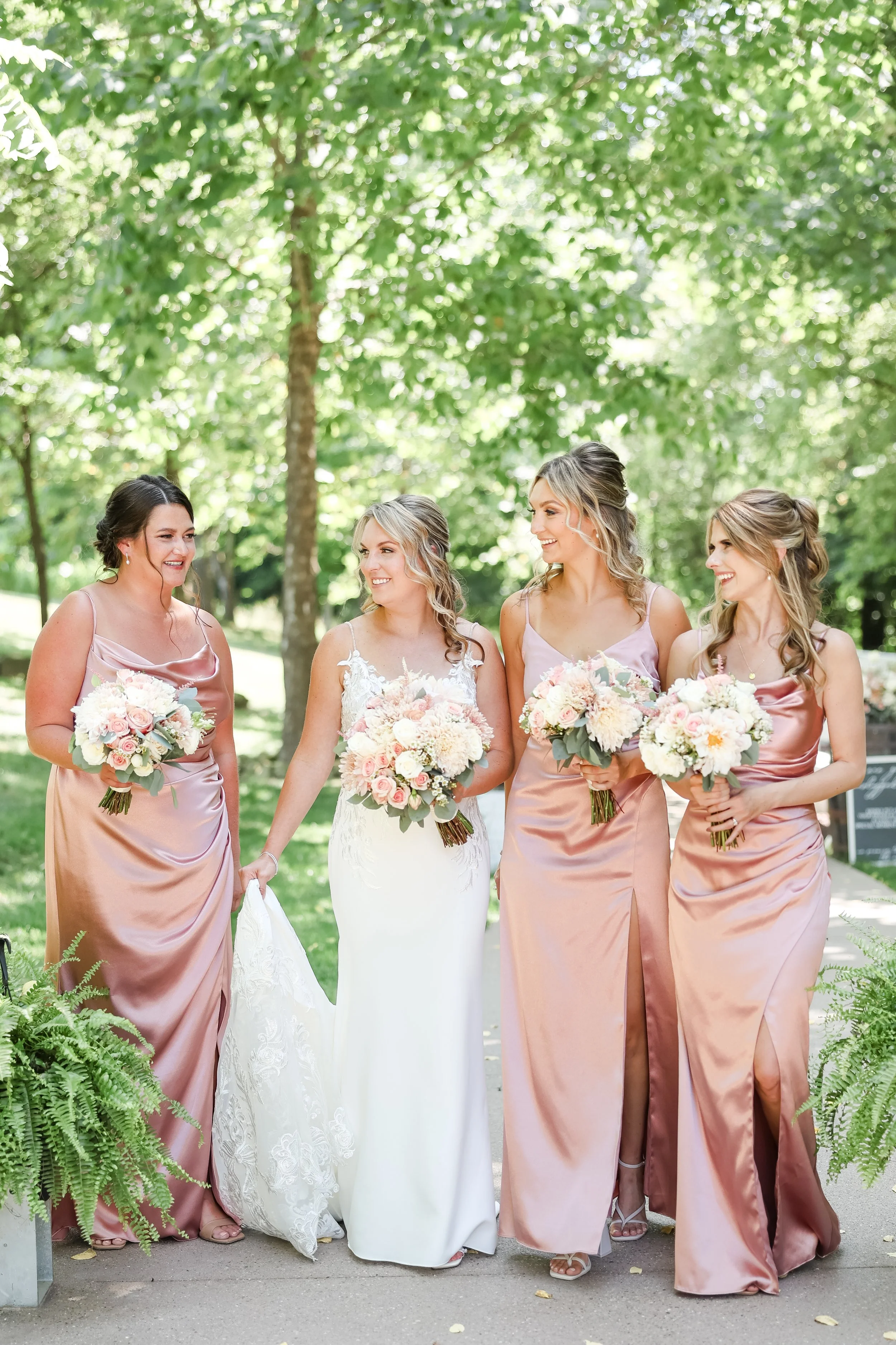 A bride and three bridesmaids walking outdoors, holding bouquets of pink and white flowers, surrounded by greenery.