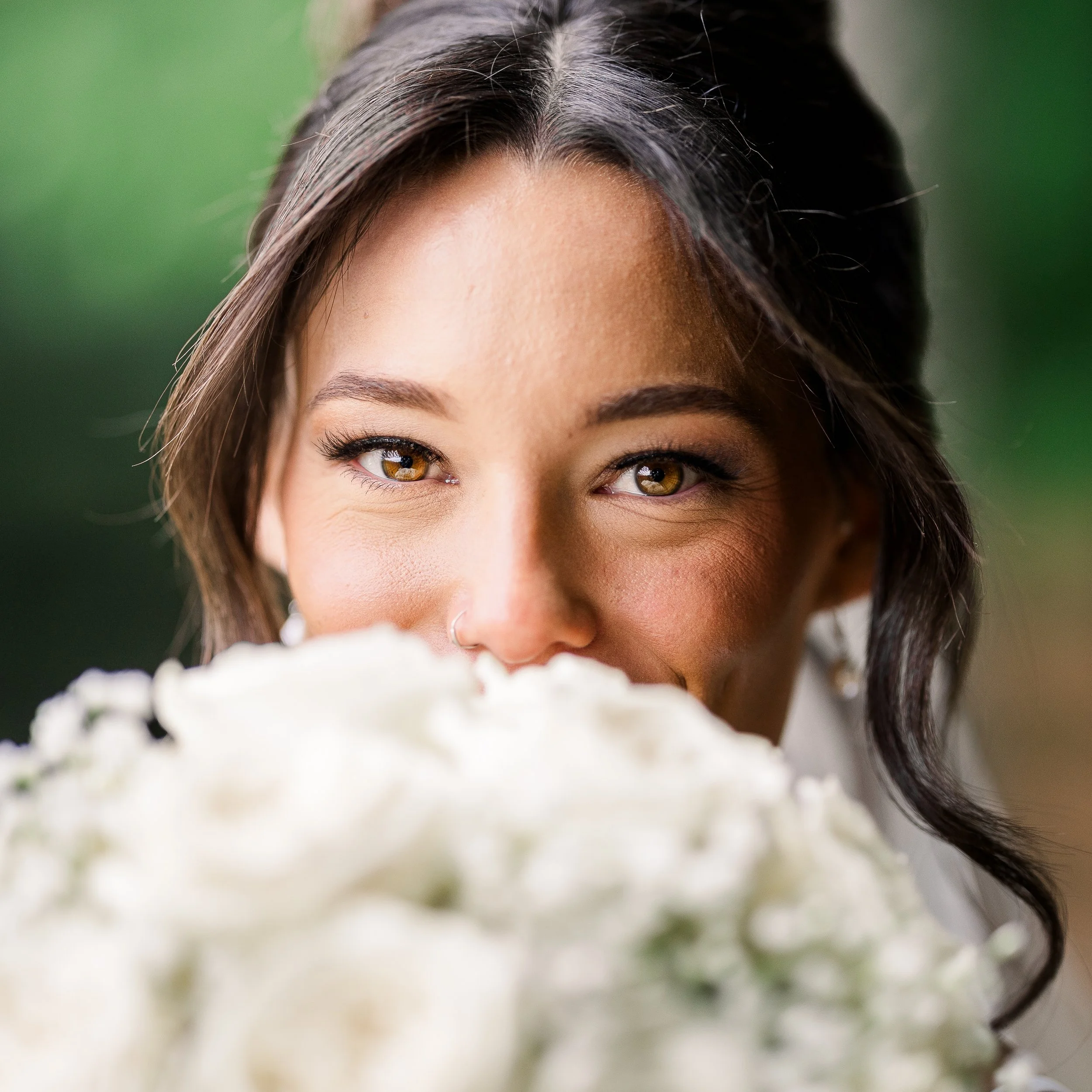 A woman with brown hair and hazel eyes peeking over a bouquet of white flowers, with a blurred green background.