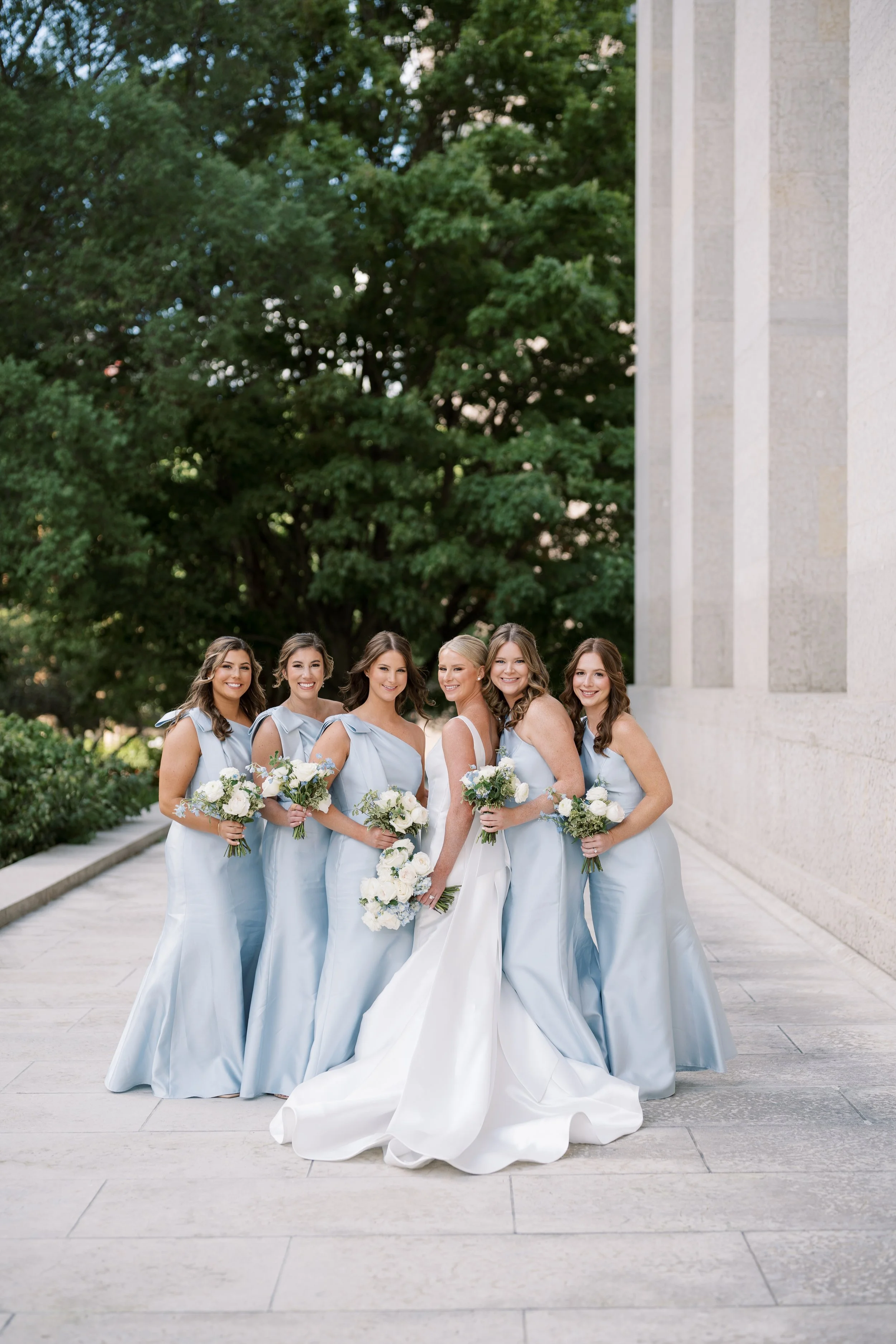 Bride and six bridesmaids in light blue dresses holding bouquets standing outdoors in front of a stone building and green trees.
