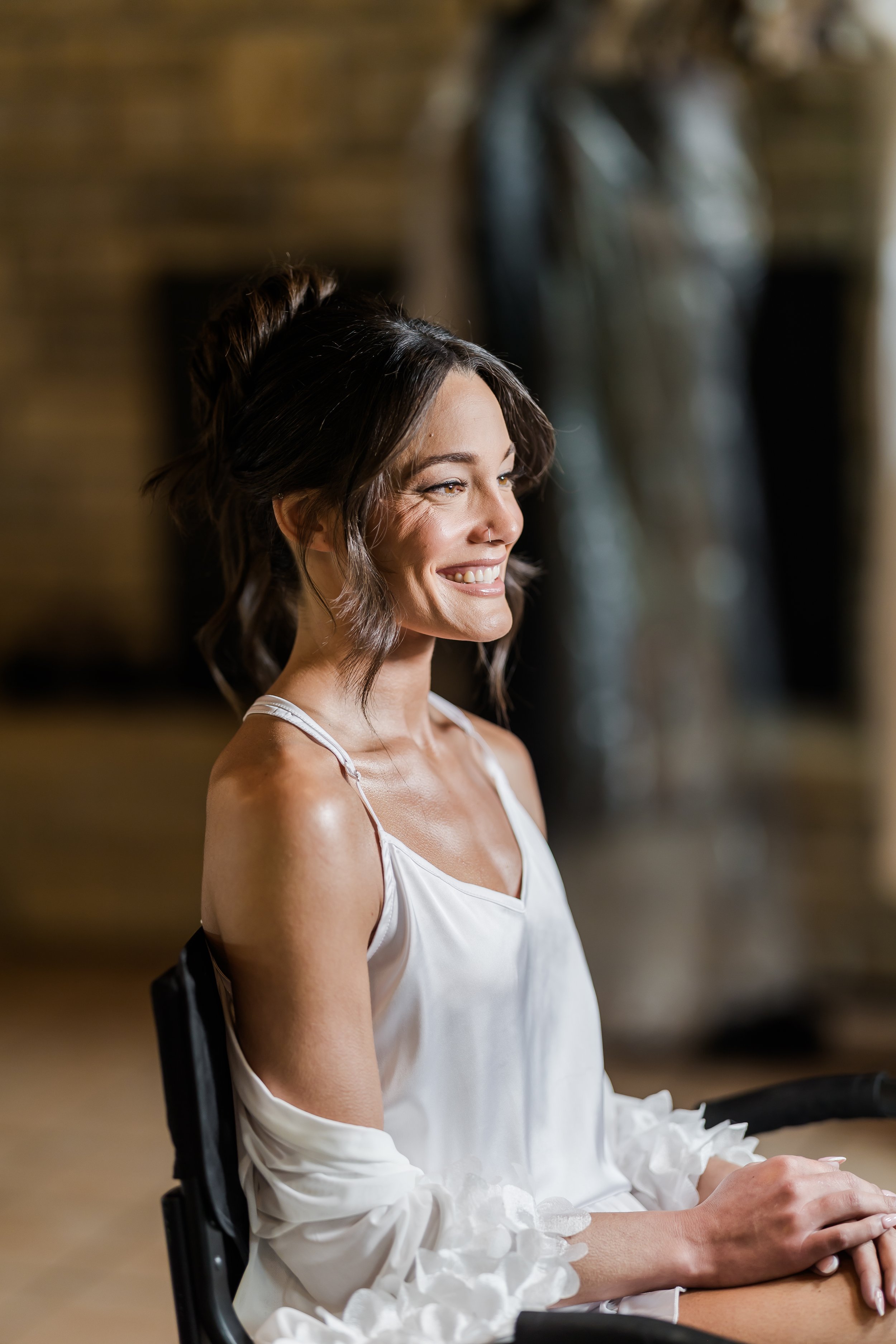 A smiling woman with short brown hair sitting in a chair, wearing a white dress with thin straps and lace details on the sleeves. The background features a brick wall and a sculpture.
