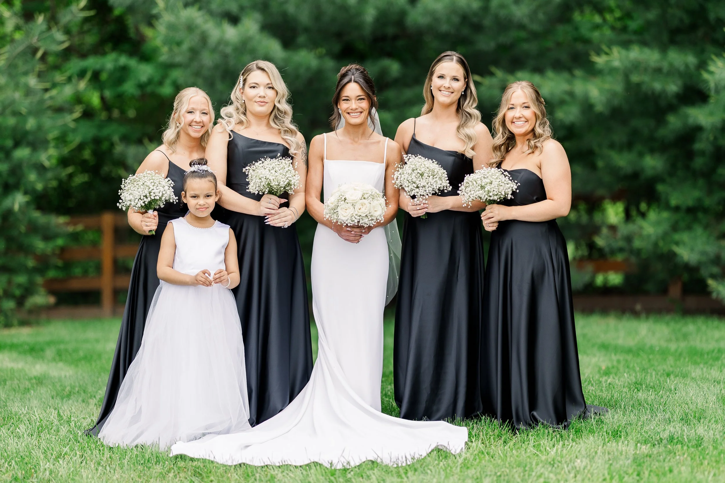 A group of women and a girl at a wedding outdoors, with the bride in a white dress holding a bouquet, surrounded by bridesmaids in black dresses and a young girl in a white dress, all smiling and holding flowers.