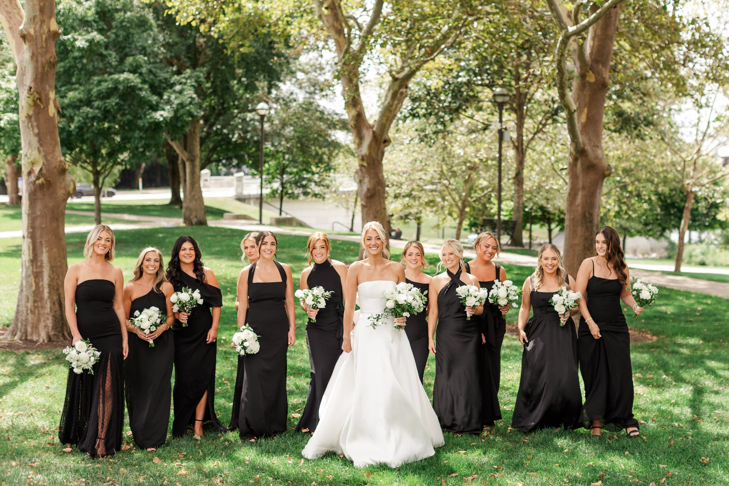 A bride and nine bridesmaids standing outdoors in a park, all holding white flower bouquets, with trees and green grass in the background.