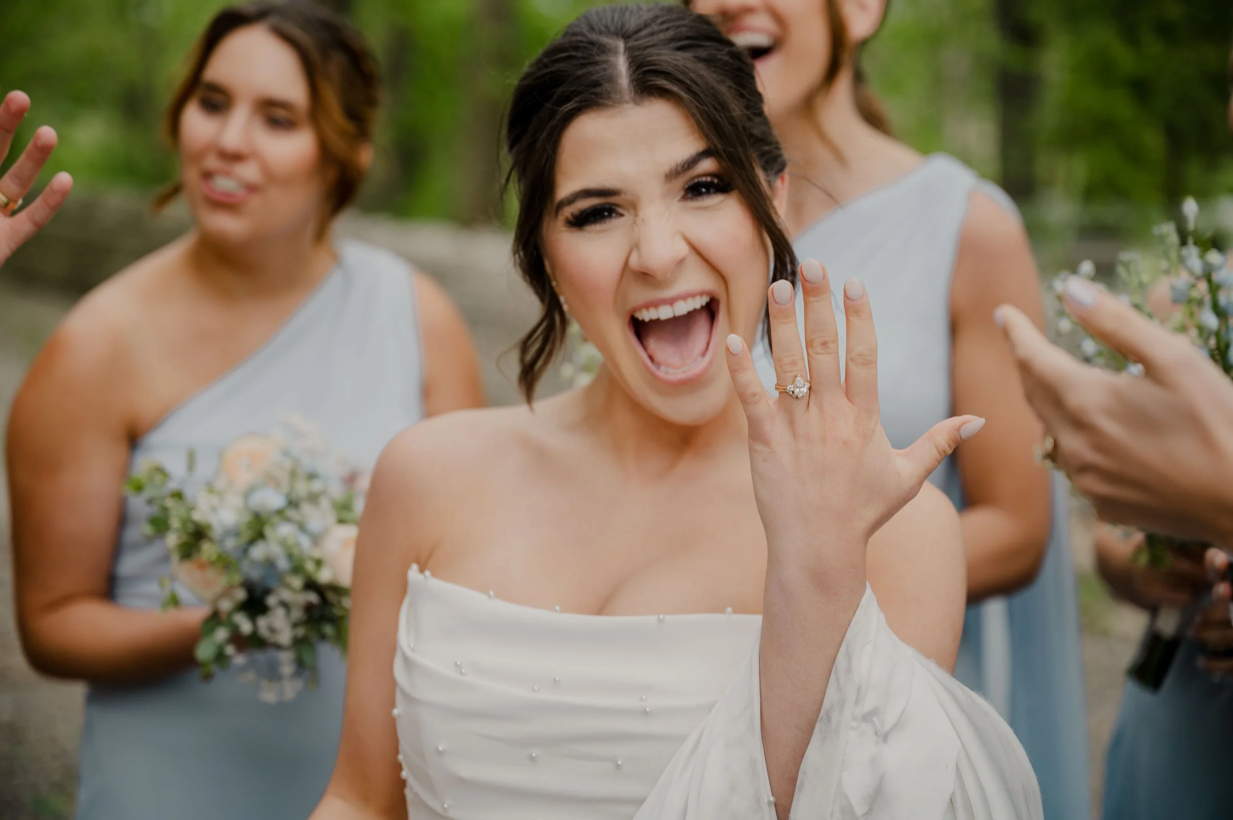 A bride showing her wedding ring and smiling, surrounded by her bridesmaids outdoors.
