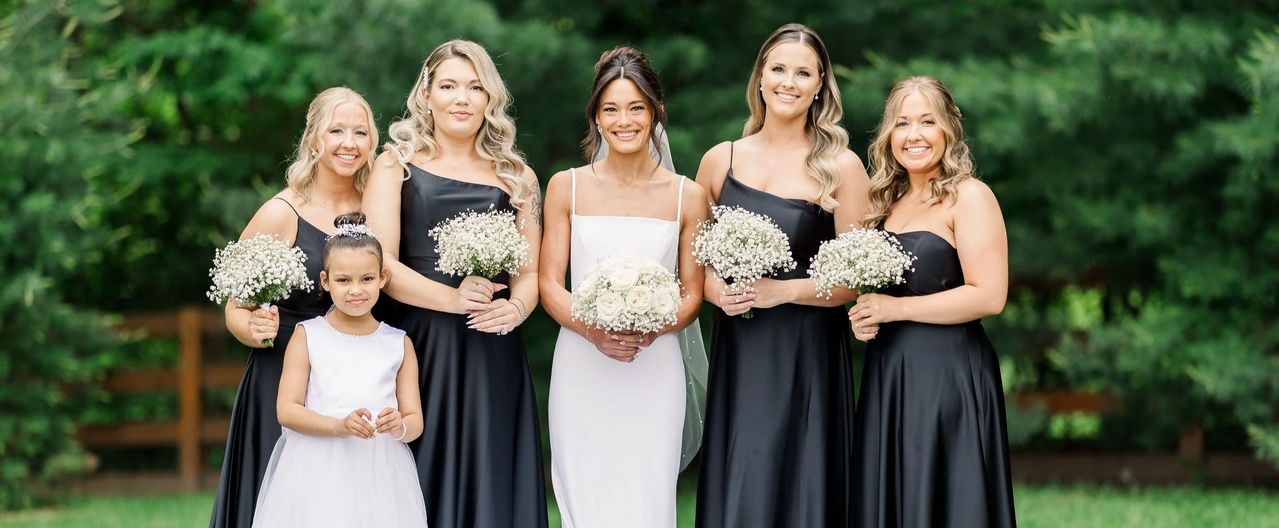 A bride in a white dress holding a bouquet of white roses stands among five women in black dresses holding white floral bouquets, outdoors with green trees in the background.