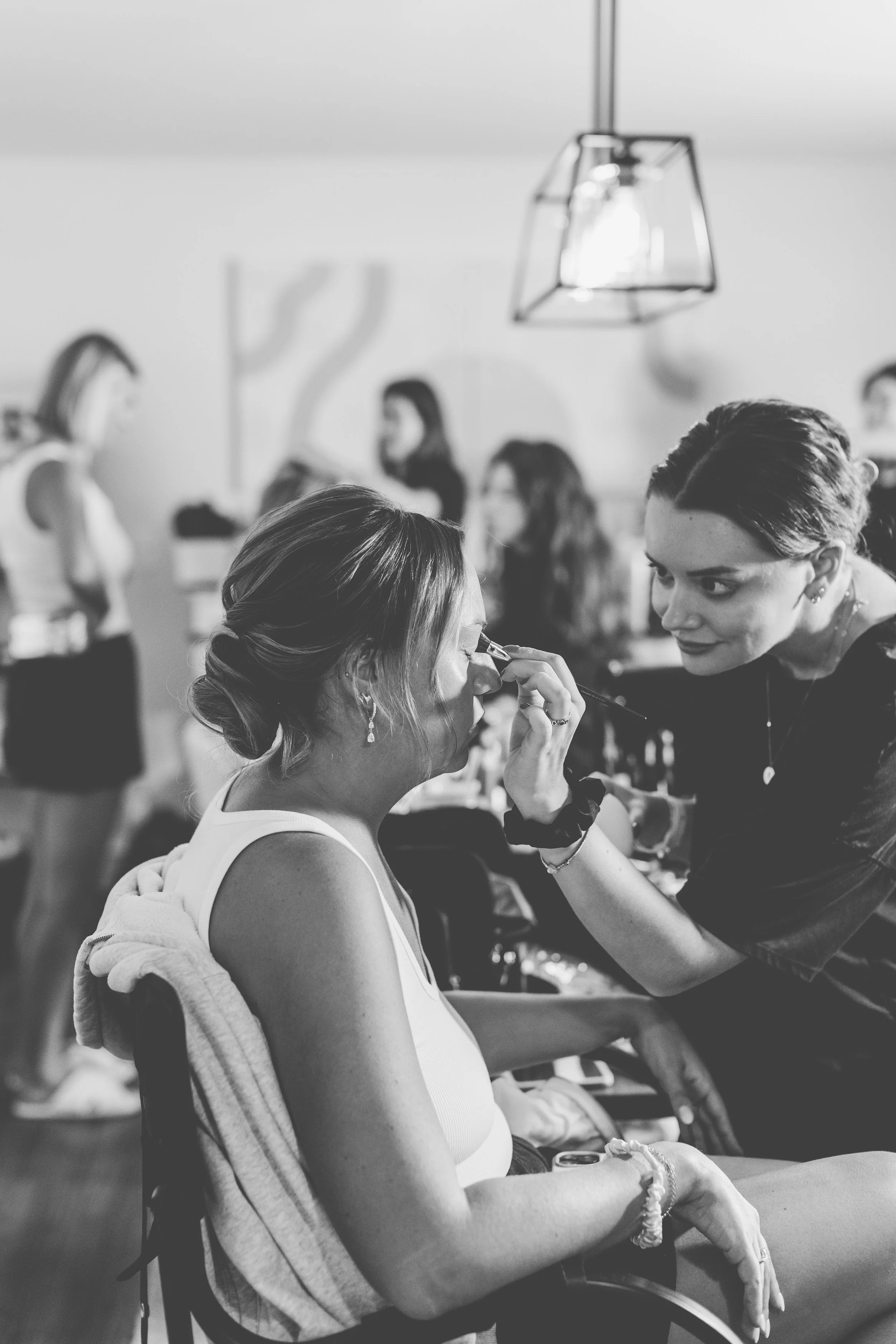 Women getting makeup applied in a busy salon with other clients and staff in the background.