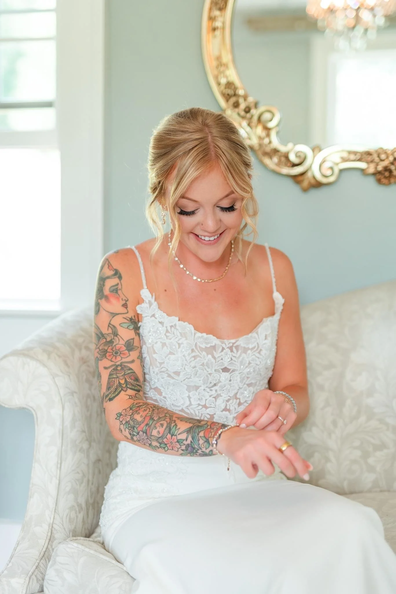 A smiling woman in a white lace dress is sitting on a cream-colored sofa, adjusting her bracelet. She has blonde hair in an updo, tattoos on her arms, and is wearing jewelry, with a large ornate mirror and window in the background.