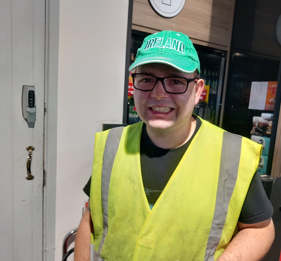 A young man wearing glasses, a green Irish cap, and a yellow reflective vest, standing indoors, smiling at the camera.