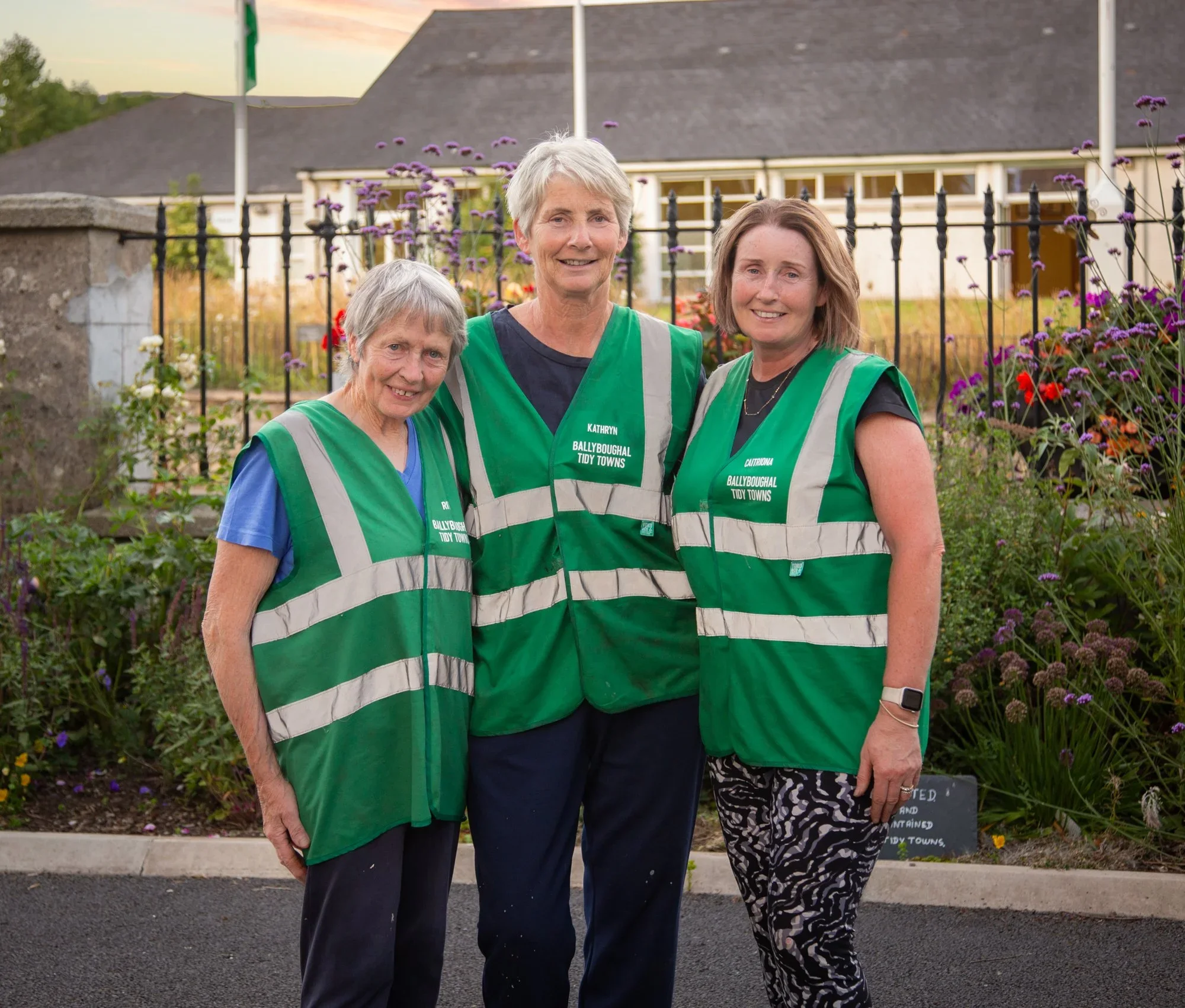 Three women wearing green safety vests standing outdoors in front of a garden with flowers, a fence, and a house in the background, smiling for the photo.