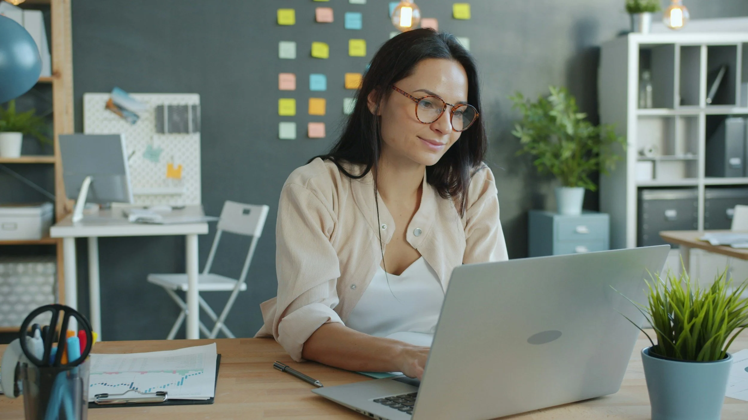 A woman with dark hair and glasses working on her laptop at a desk in an office environment with plants and sticky notes on the wall.
