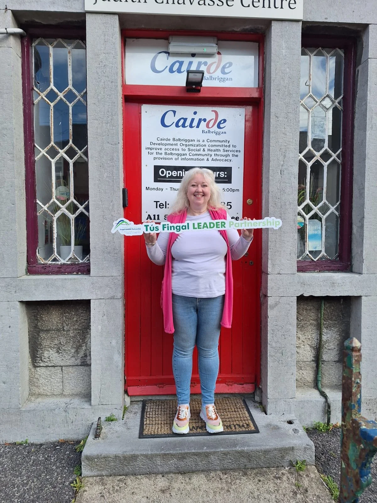 A smiling woman standing outside a red door of a community center, holding a sign that reads 'Tús Fíngal LEADER Partnership.' The entrance has two windows with decorative metal grilles, and a sign above the door reads 'Cairde Balbriggan.'