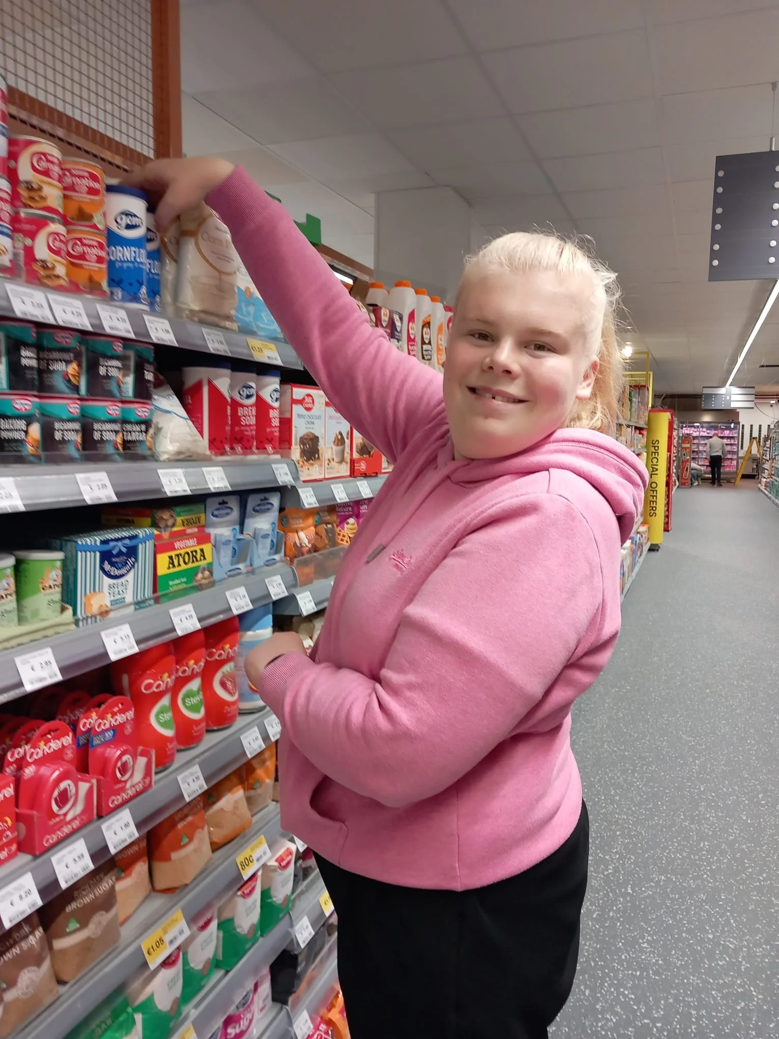 A young girl with blonde hair wearing a pink hoodie and black pants reaching for a product on a grocery store shelf, smiling at the camera.