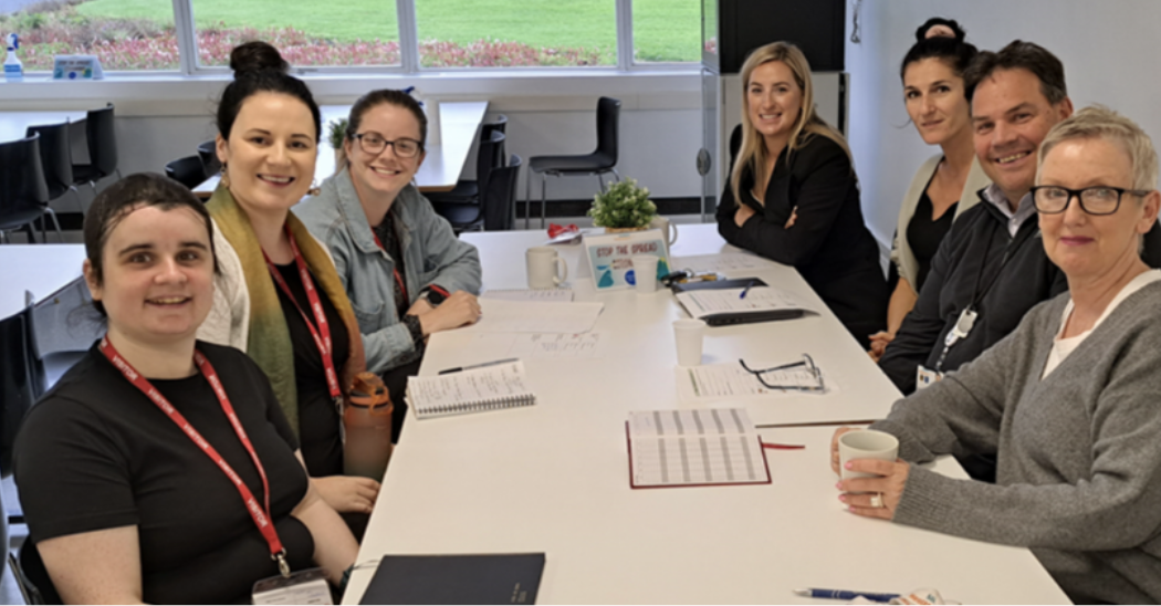 Seven people sitting around a conference table in a well-lit room with large windows. They are smiling and appear to be in a meeting or discussion, with papers, notebooks, and cups on the table.