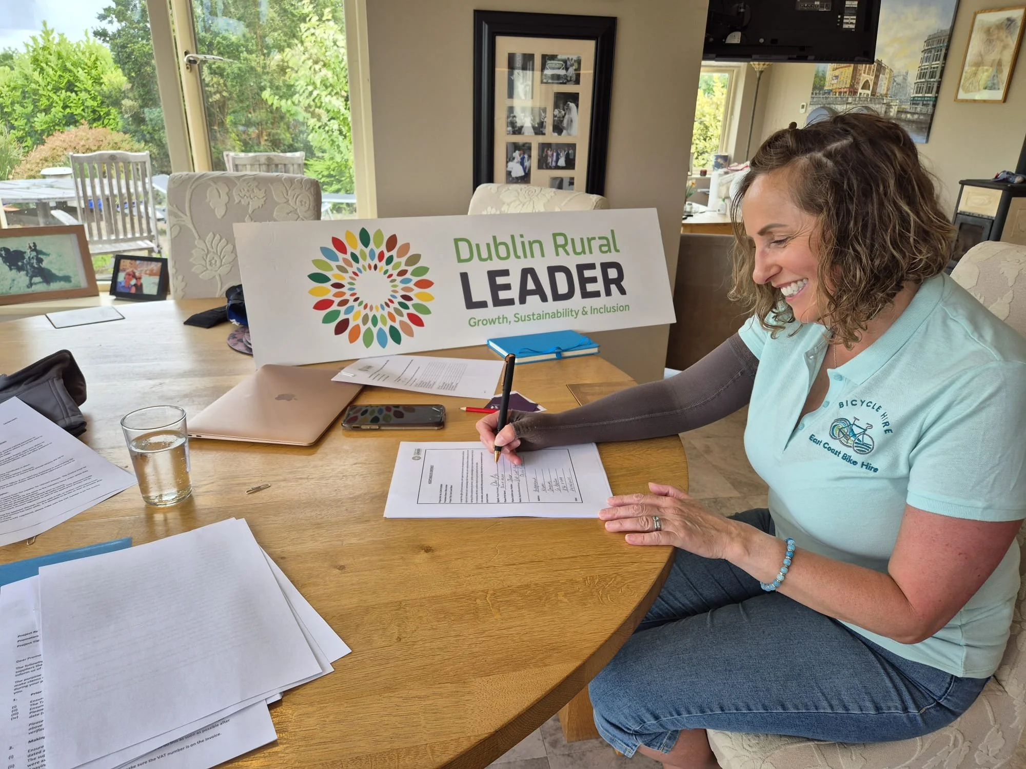 A woman sits at a dining table, signing a document, with a glass of water, laptop, and phone on the table. There is a sign in the background that reads 'Dublin Rural Leader - Growth, Sustainability & Inclusion'. She is wearing a light blue polo shirt with a logo that says ‘Bicycle Hire - East Coast Bike Hire’. The room has large windows with trees outside, and photos on the wall.