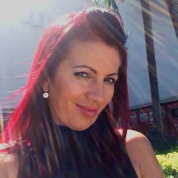 A woman with red hair smiling outdoors with sunlight on her face and a palm tree in the background.