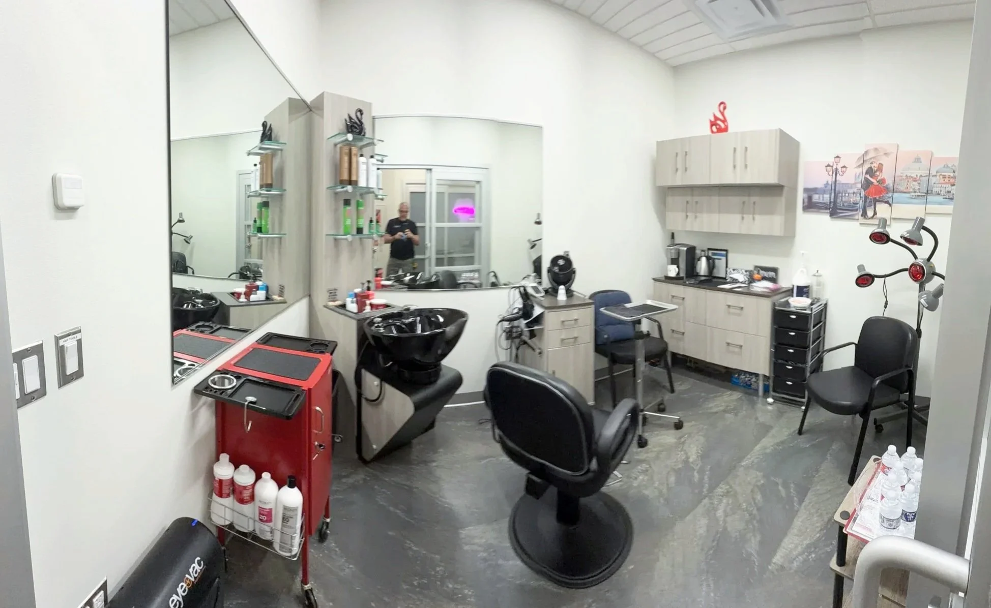A hair salon or beauty treatment room with chairs, mirrors, and hair styling equipment, including a wash sink, storage cabinets, and a person taking a photo in the mirror.