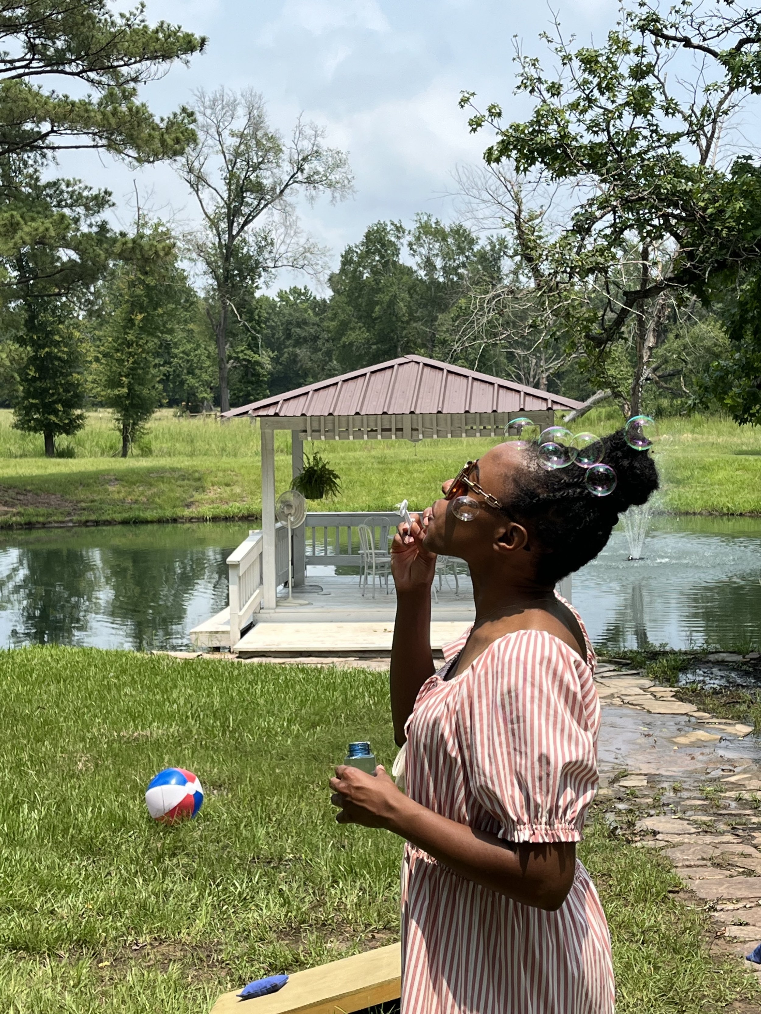 A woman with glasses and curly hair in an updo, wearing a striped dress, stands near a pond, blowing bubbles. A beach ball is on the grass nearby, and a small wooden pier extends into the pond with a gazebo in the background surrounded by lush green 