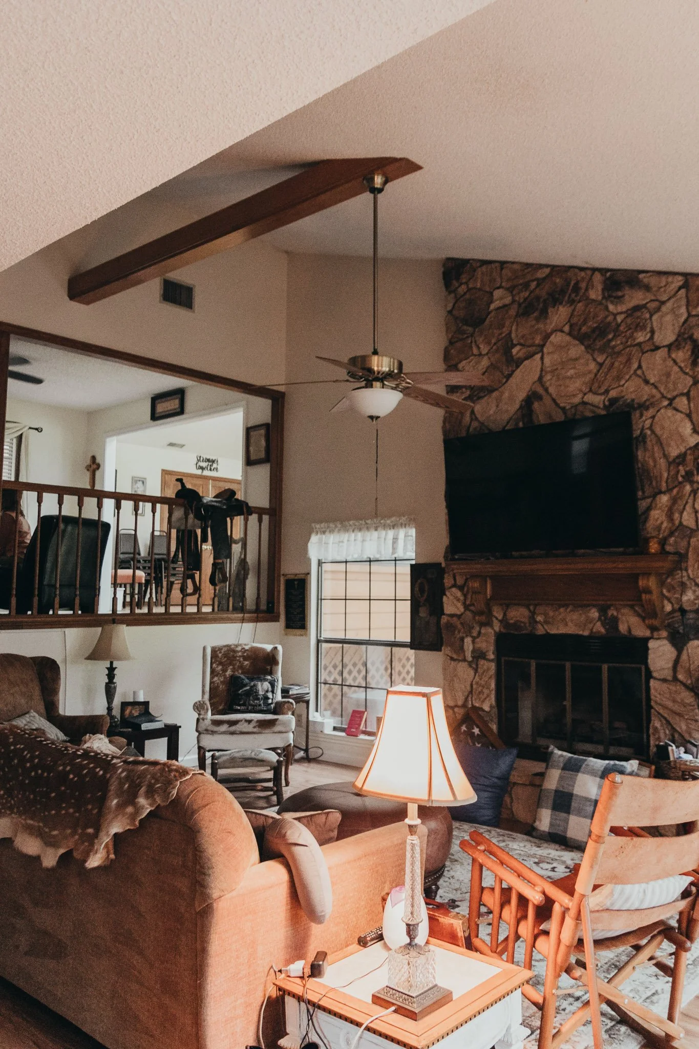 Living room with a stone fireplace, flat-screen TV, ceiling fan, table lamp, armchair, sofa, and wooden chairs, with a staircase and dining area visible in the background.