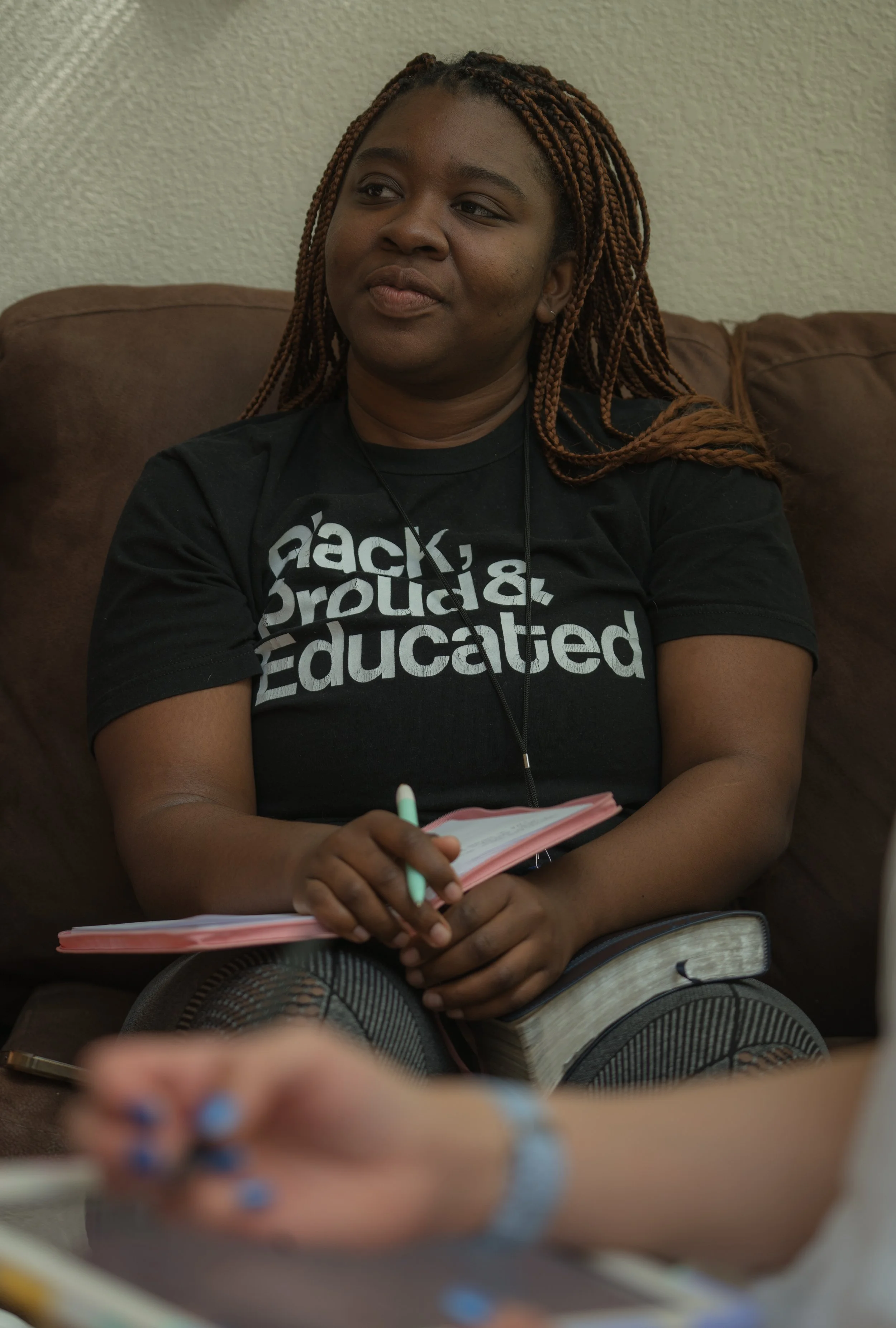 A woman with braids sitting on a brown couch, holding a notebook and pen, wearing a black T-shirt that says 'Black, Proud & Educated'.