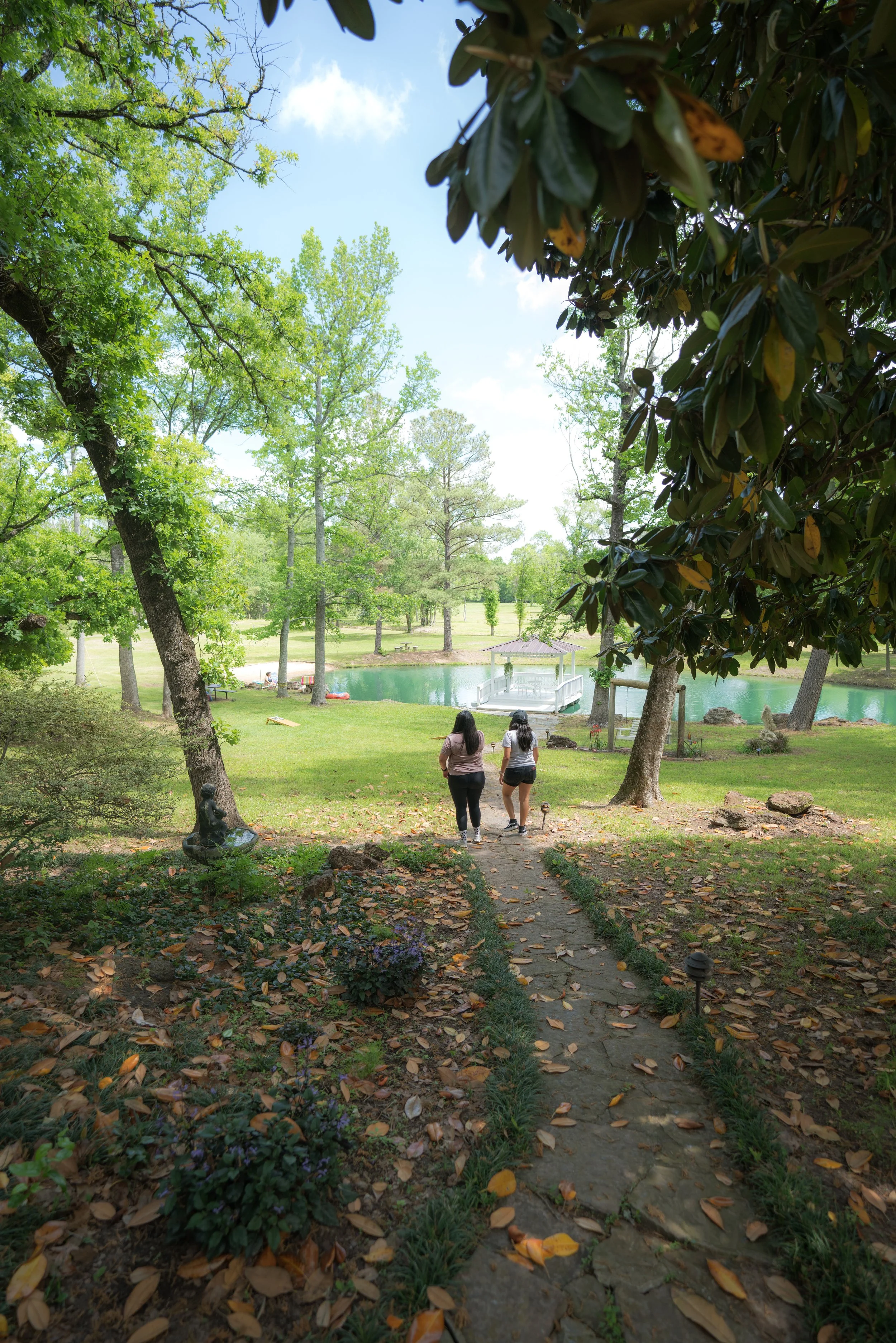 Two women walking on a stone pathway in a park with green trees, a pond, and a gazebo in the background.