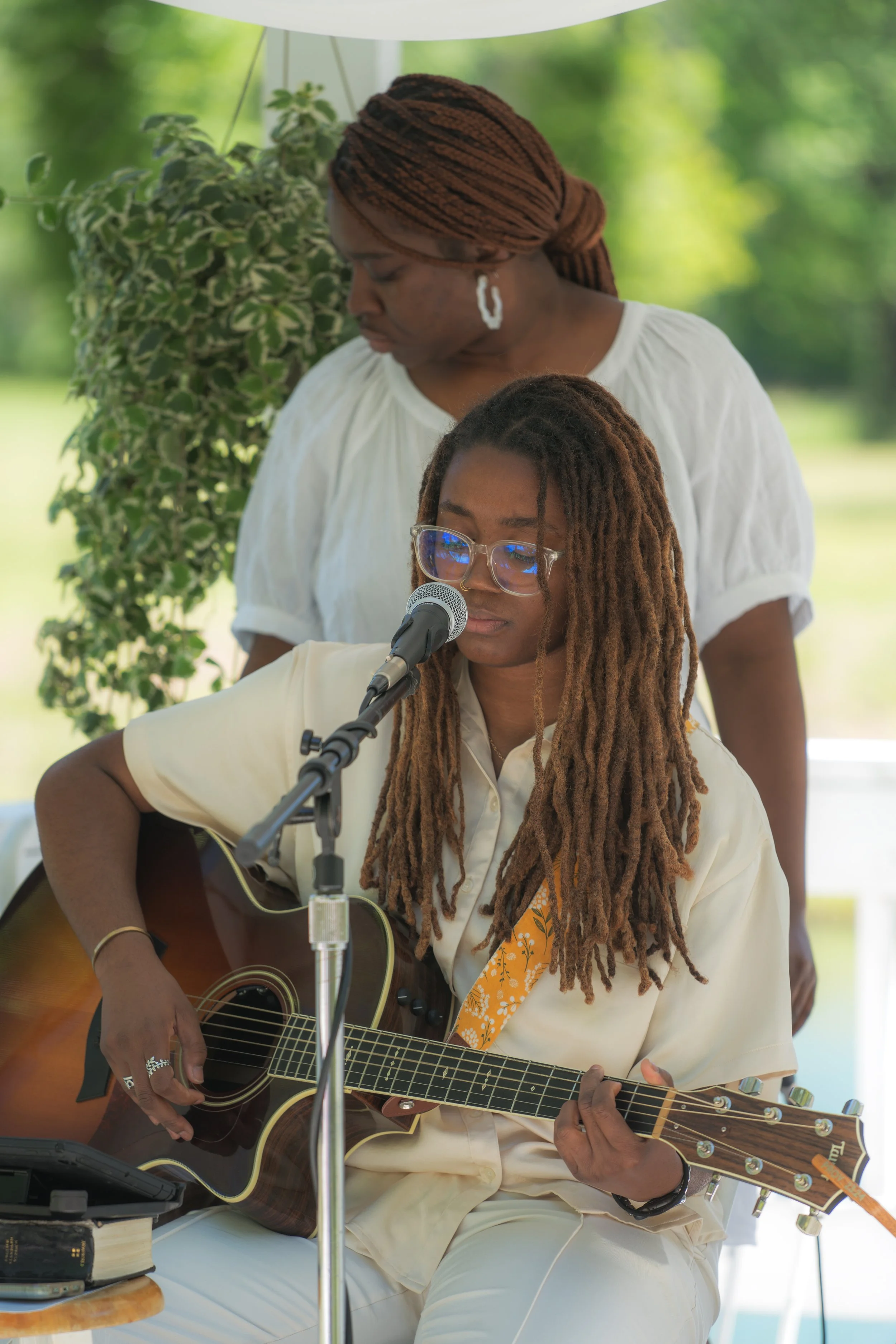 A woman with glasses and dreadlocks playing an acoustic guitar and singing into a microphone, with another woman standing behind her, outdoors in a park-like setting.