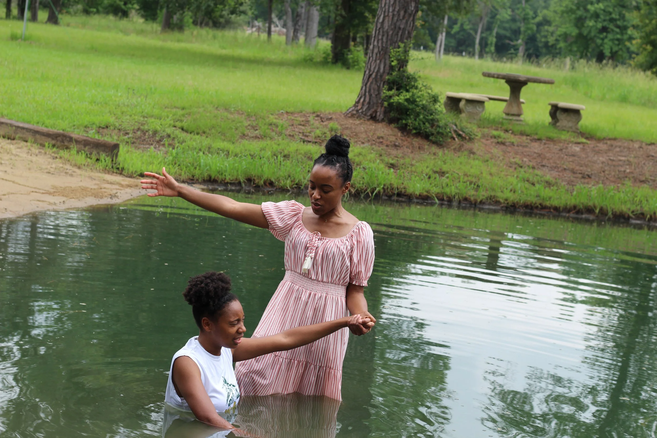 Two women are in a pond, one standing in the water and the other sitting, holding her hand. The standing woman is wearing a pink and white striped dress, and the sitting woman is wearing a white sleeveless top. The scene is outdoors with grassy areas