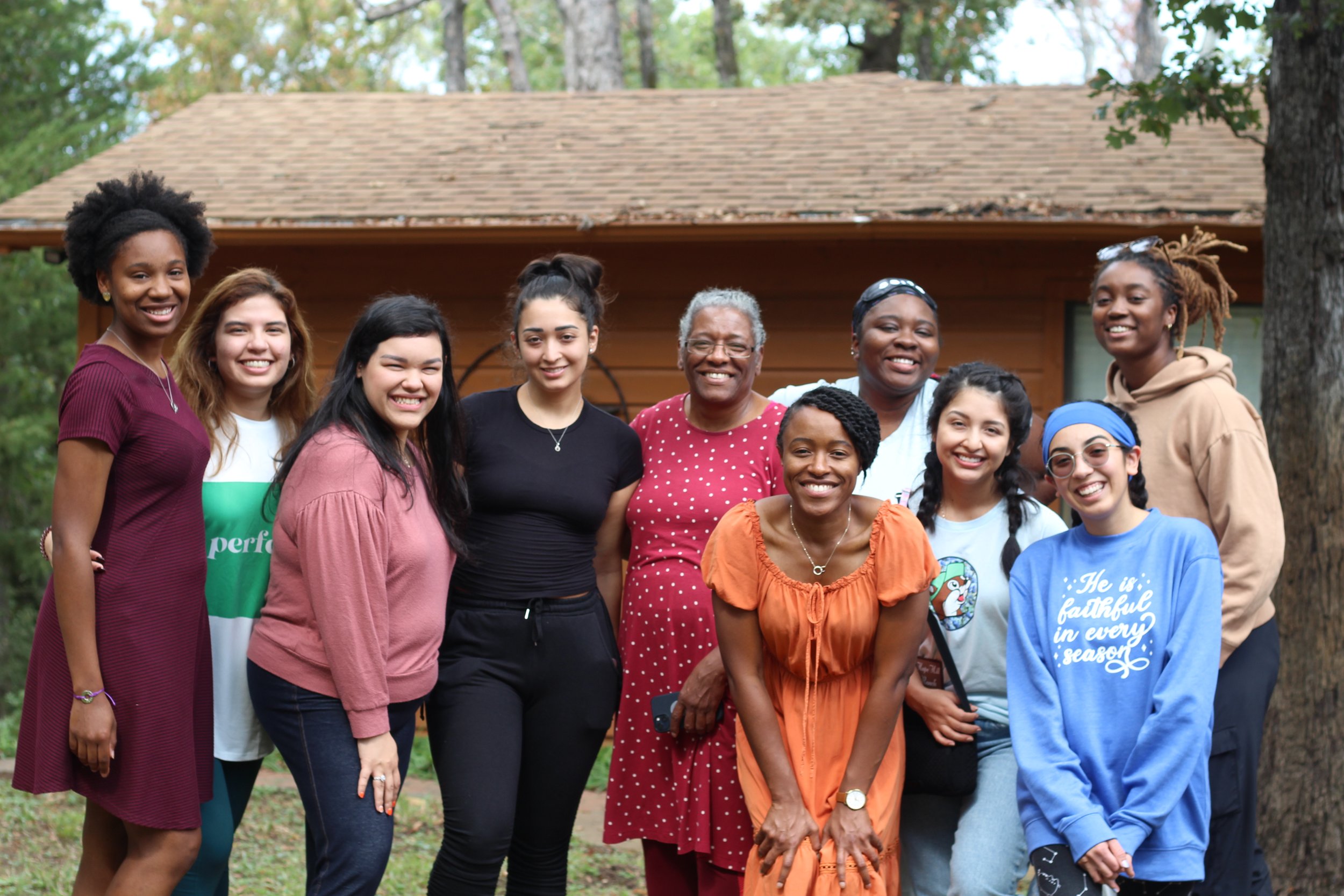 Group of diverse women smiling outdoors in front of a wooden house and trees.