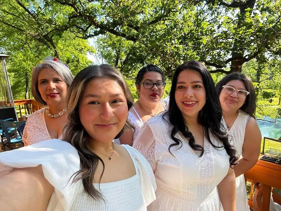 Five women smiling outdoors, standing under a tree with green leaves, in a park or garden setting.