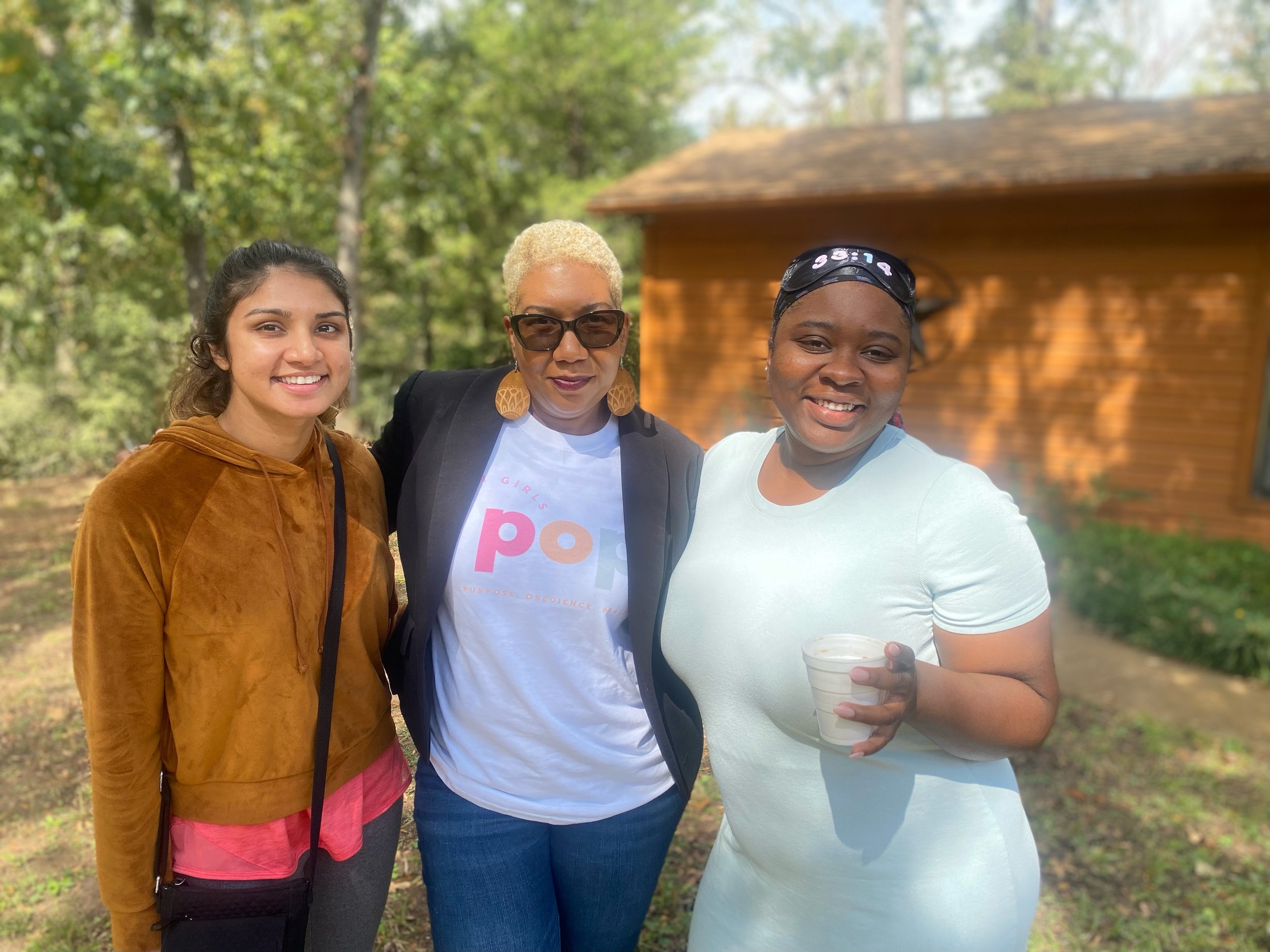 Three women standing outdoors in front of a wooden building and trees, smiling at the camera. One woman is holding a small cup.
