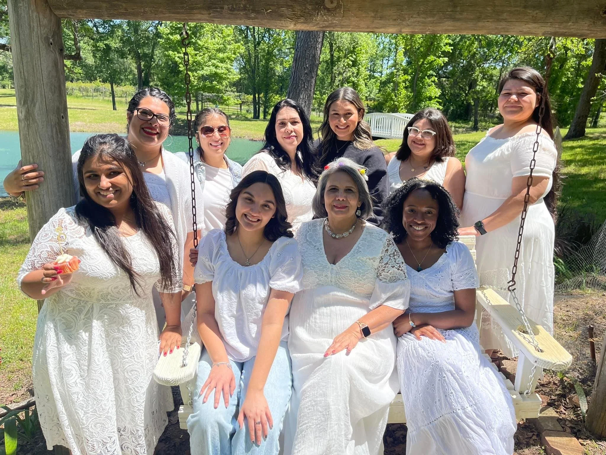 A group of twelve women gathered outdoors, sitting and standing around a white swing on a sunny day, with greenery and trees in the background, all dressed in white clothing.